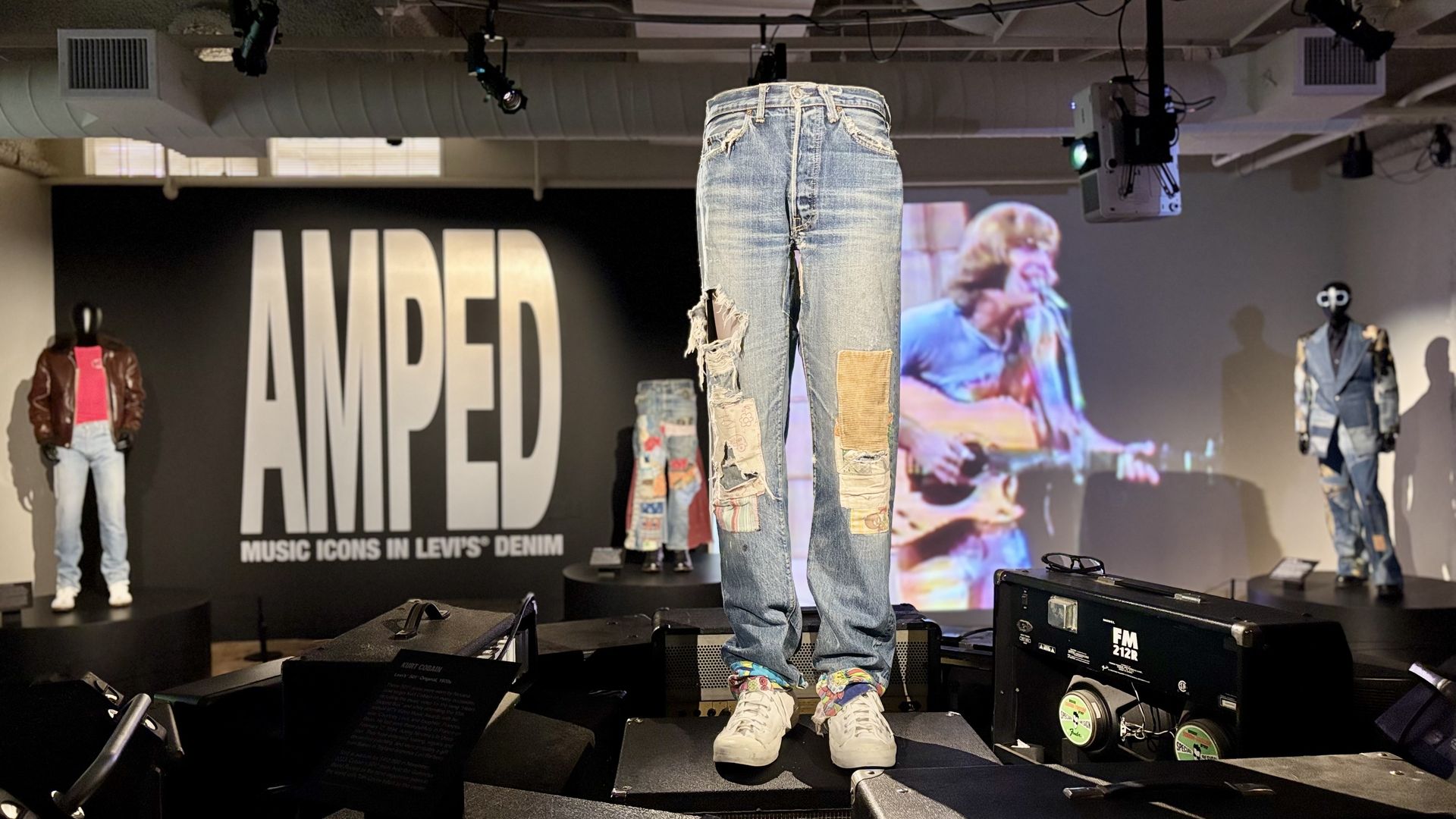Display of Kurt Cobain's patched, ripped Levi's denim jeans and white shoes at the AMPED exhibit featuring music icons, with a blurred video of a guitarist in the background. 