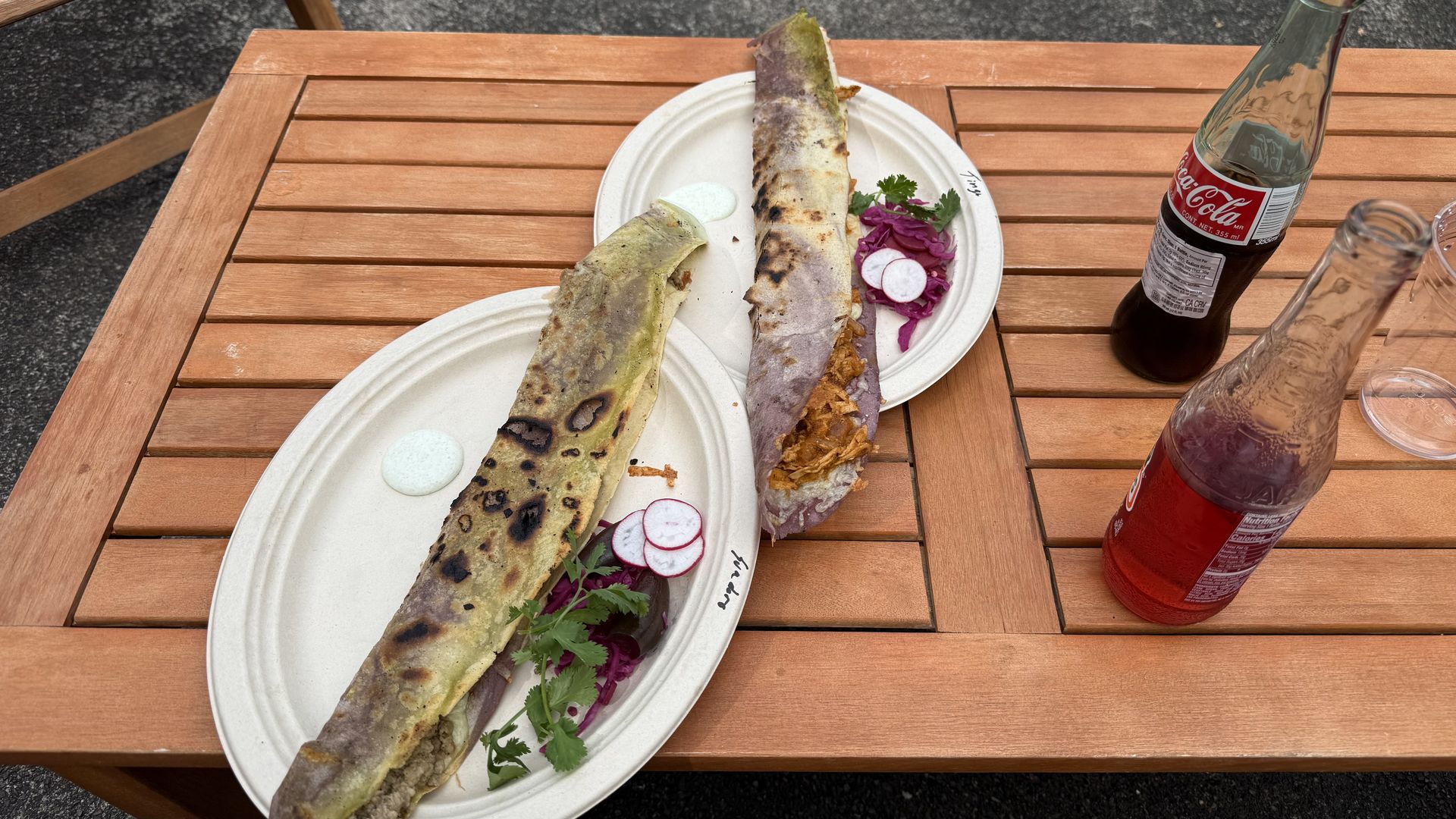 Two long grilled quesadillas on white plates with purple cabbage, radish slices, and cilantro on a wooden table with glass bottles of Coca-Cola and red soda.