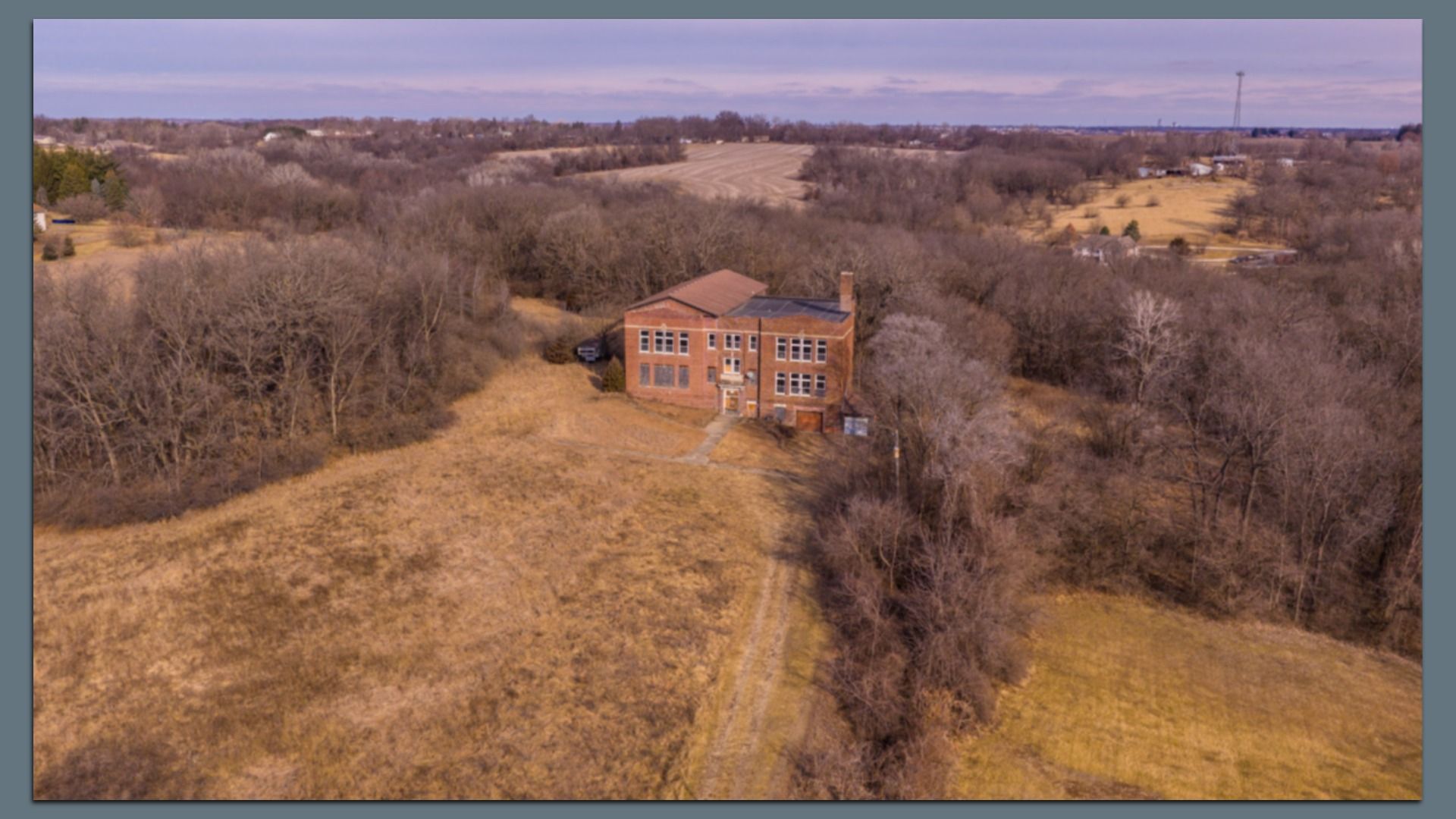 Drone view of a red brick two-story house amid leafless trees and brown fields. A dirt driveway leads to the front; a car sits behind the house, with farmland and a tower on the horizon.