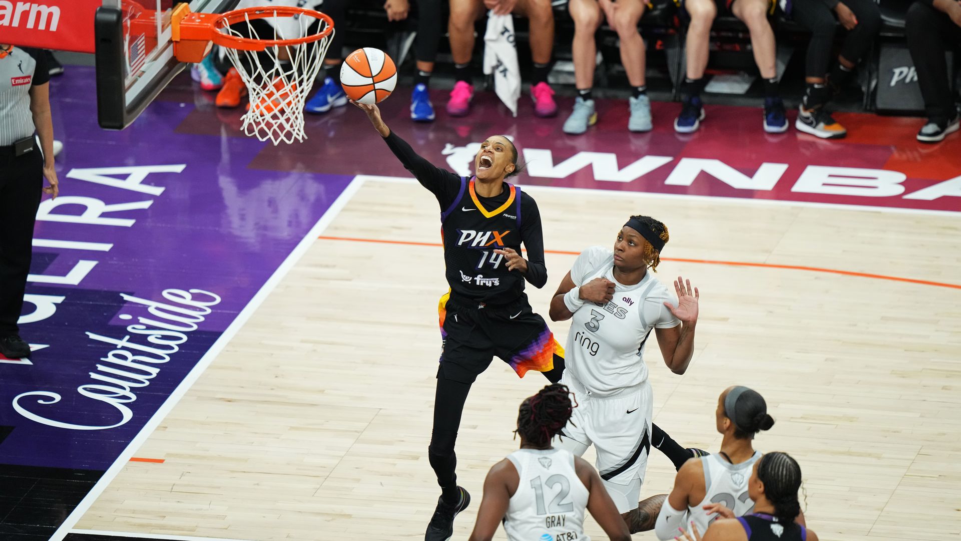 WNBA player in black PHX jersey #14 leaps for a layup while three opponents in white jerseys defend near the basket on a purple and natural wood court with 'PHX Courtside' text.
