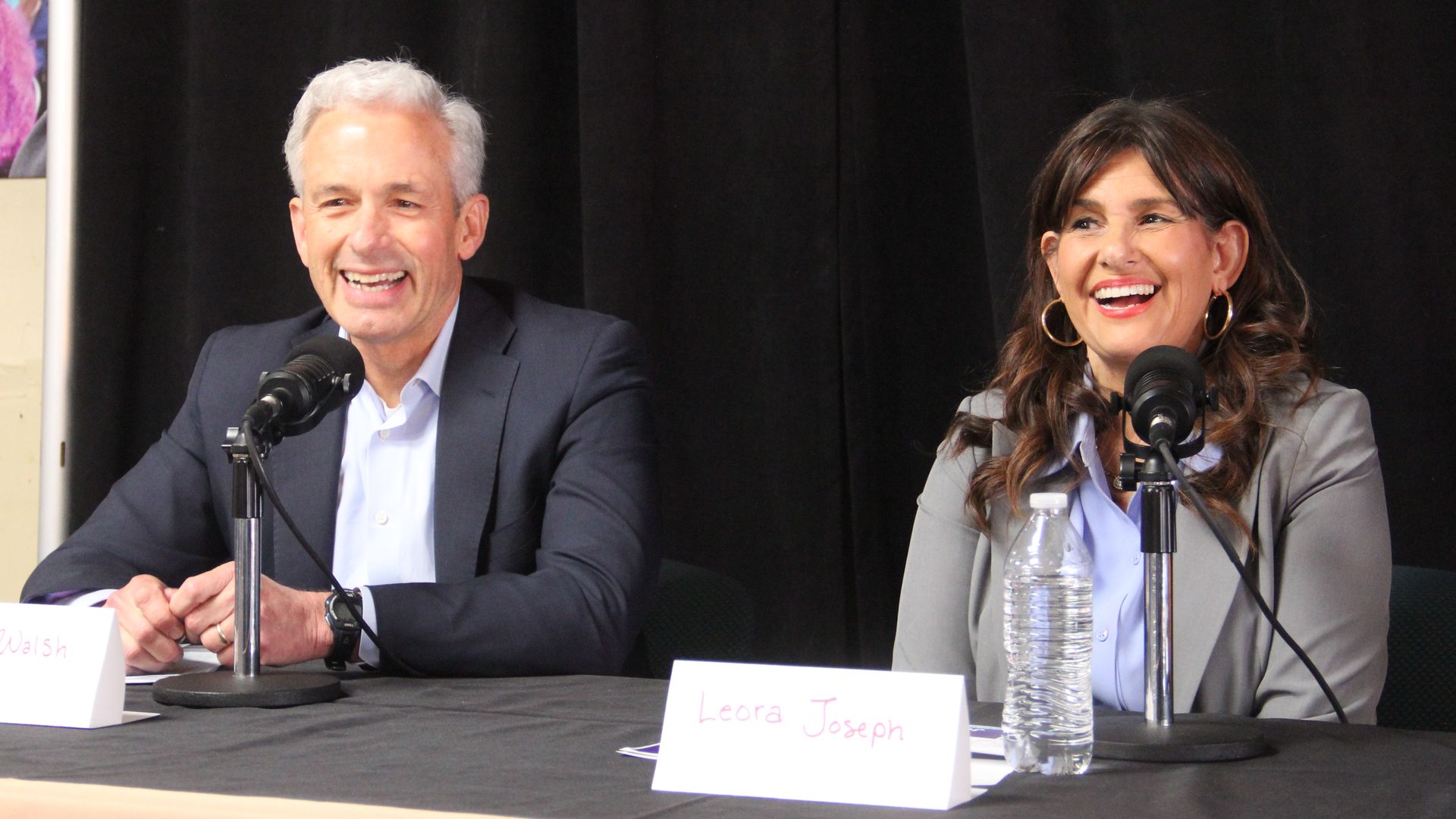 A man in a navy blazer and light blue shirt laughs while seated next to a woman in a gray blazer and light blue shirt, who is laughing as well.