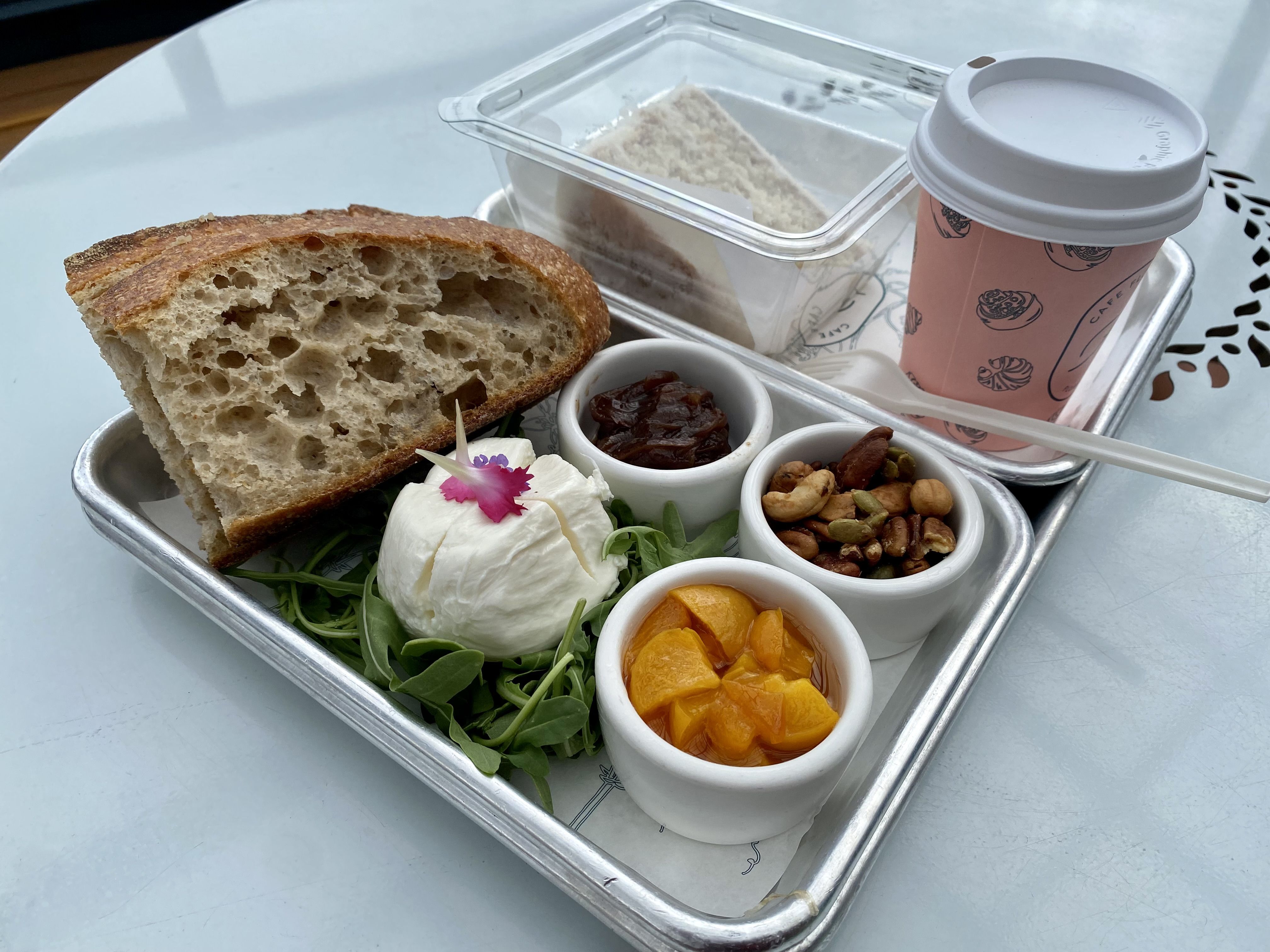 A metal tray with slices of bread, a ball of burrata and various compotes, plus a coffee cup and a clear box with a slice of cake inside.