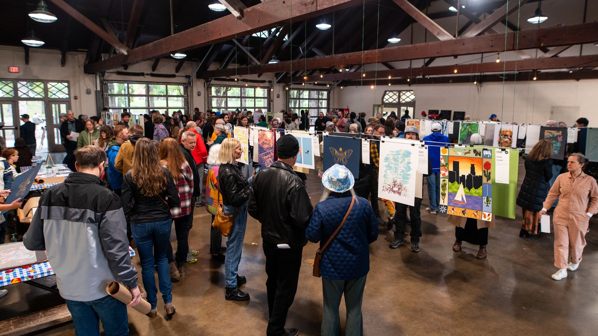Crowded indoor art show in a large hall with exposed wooden beams. People of diverse ages view colorful paintings and prints hanging from wires. Natural light filters through large windows.