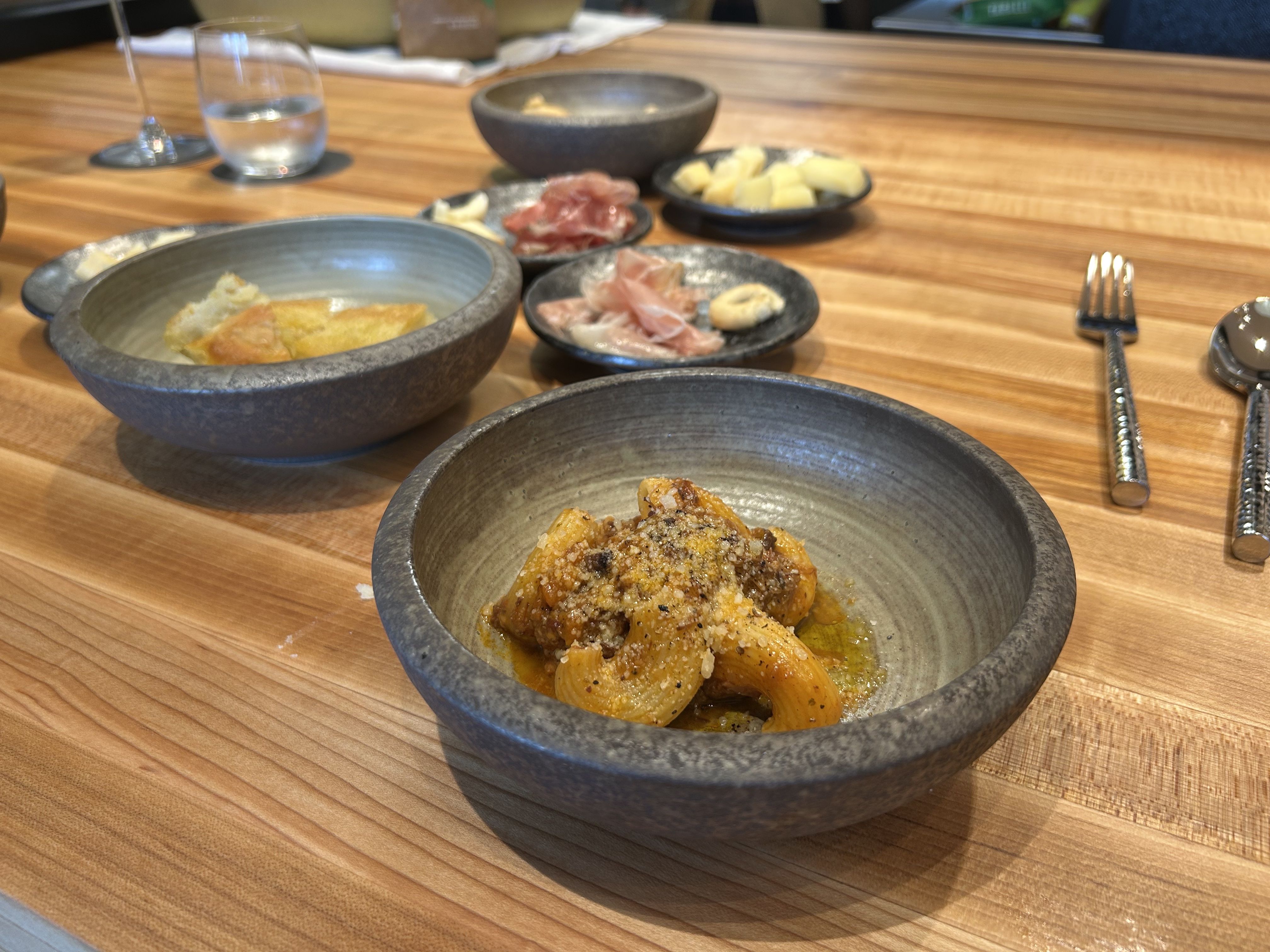 Close-up of a wooden table set with gray ceramic bowls holding various Italian dishes, including pasta with meat sauce, sliced cured meats, cheese cubes, and bread, with utensils and a glass of water nearby.