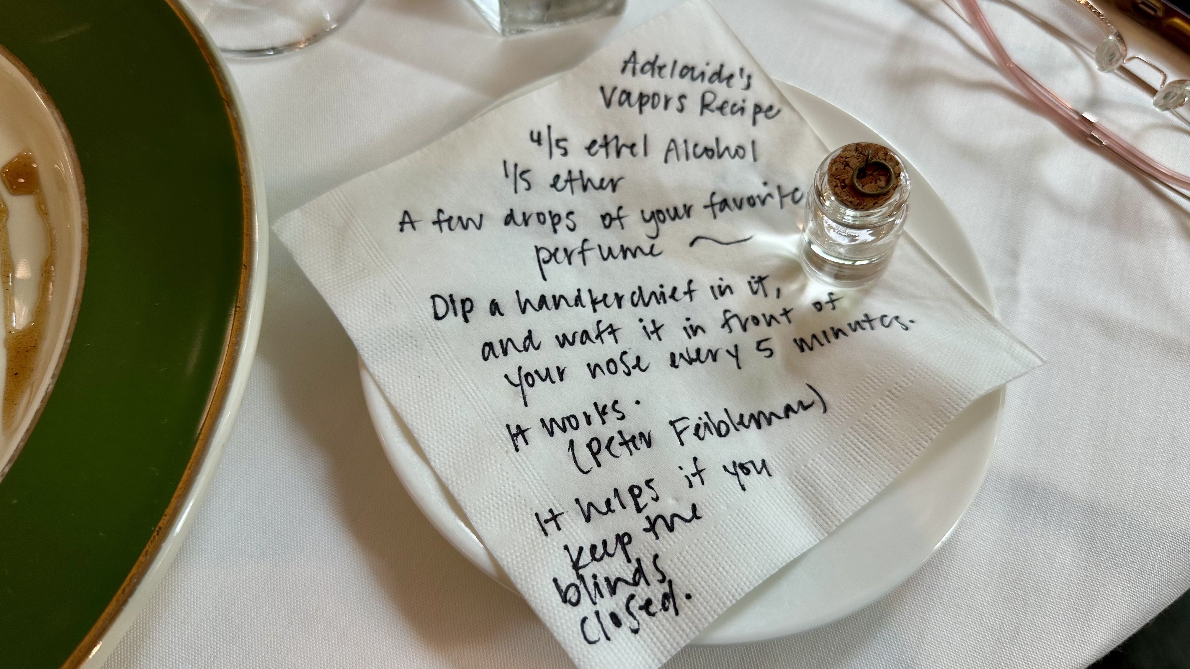 Handwritten napkin with a perfume recipe, listing alcohol and scent drops; corked glass vial sits on a white plate beside a green-rimmed dish and pink glasses on a white tablecloth.