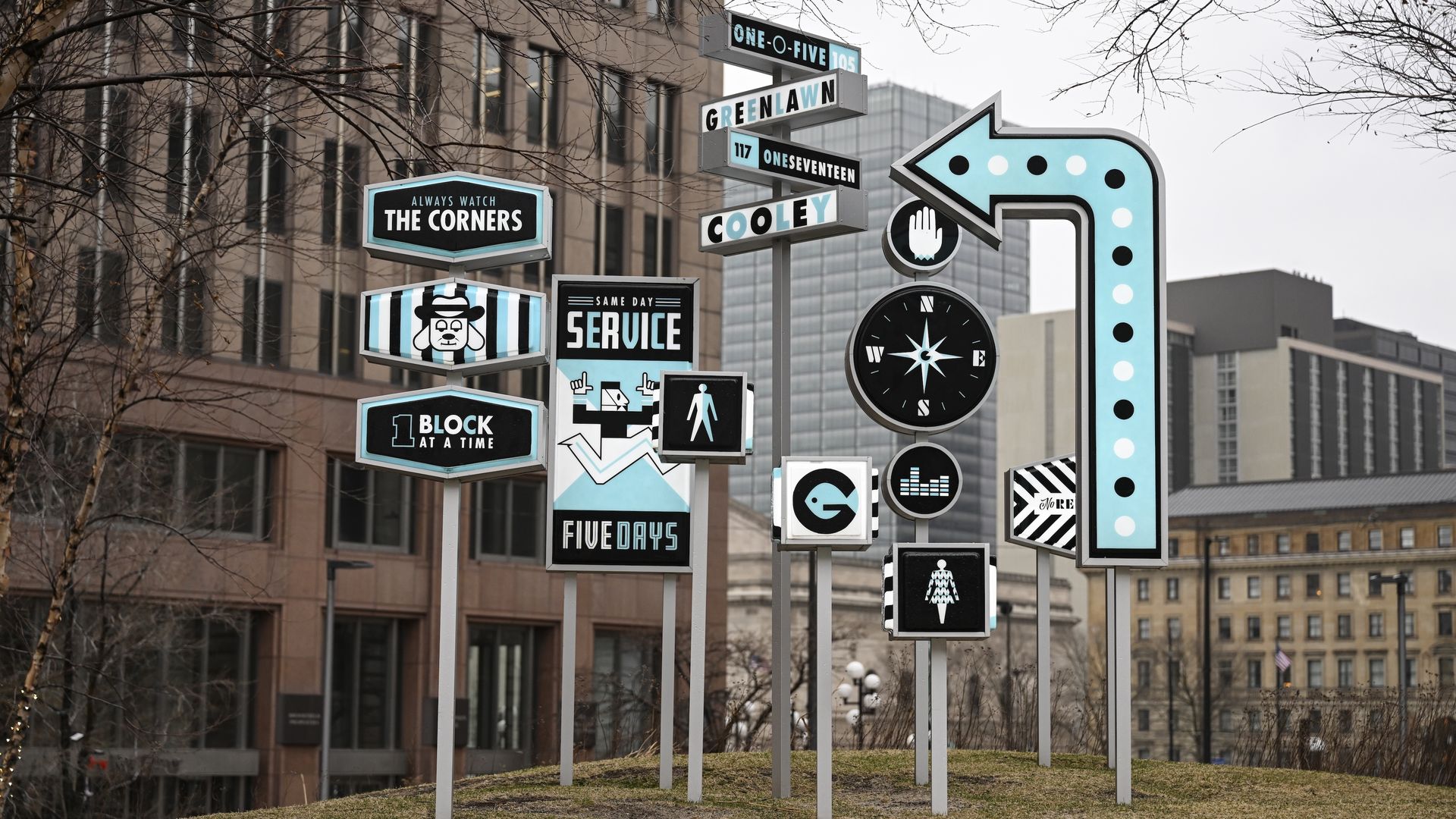 Artistic black, white, and light blue street signs clustered on poles in urban plaza, featuring arrows, pedestrian symbols, compass, and texts like \