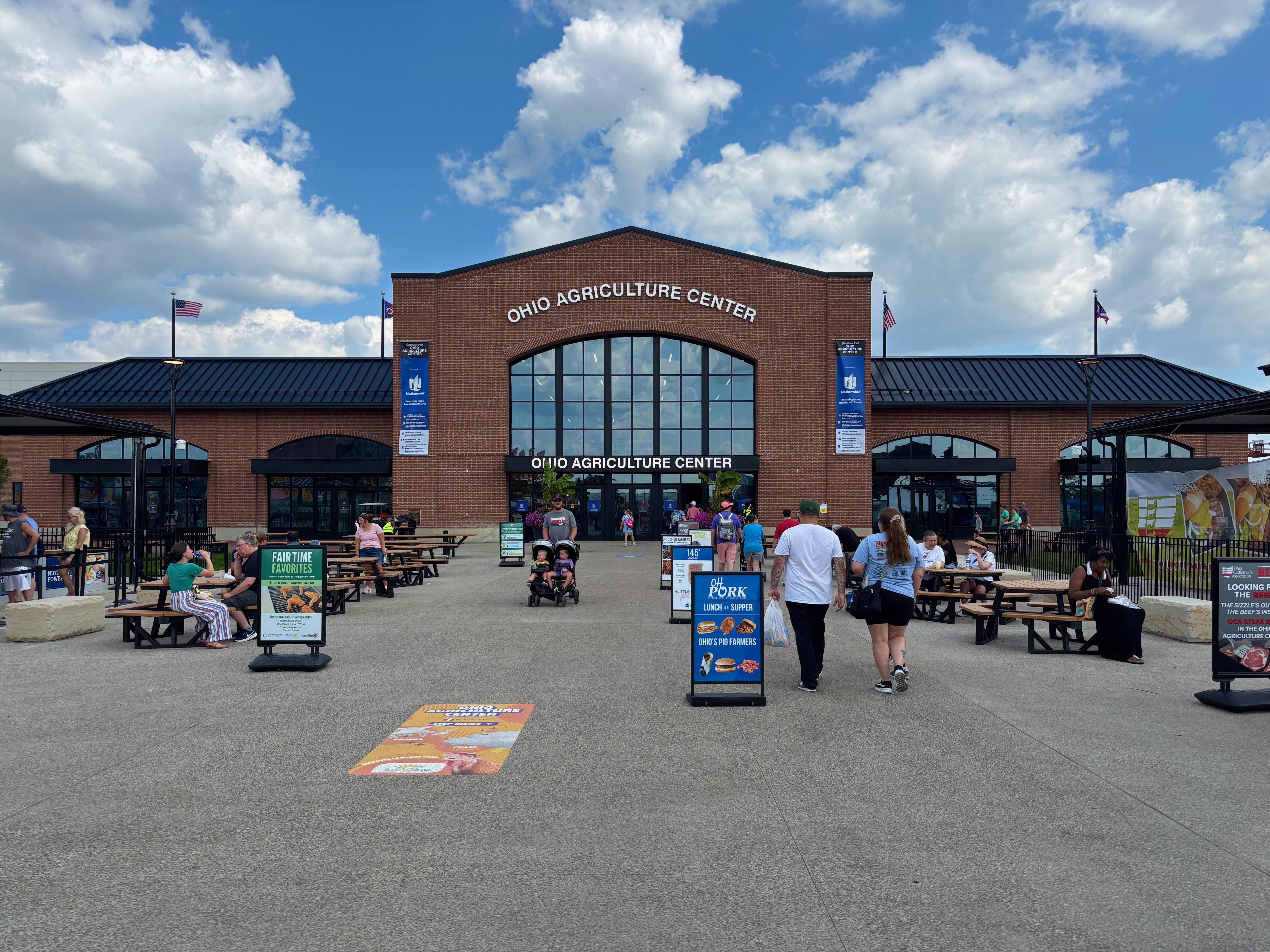 Brick Ohio Agriculture Center building with a large arched glass entrance under a bright blue sky and fluffy clouds. Outdoor tables, banners, and people walking toward the entrance.