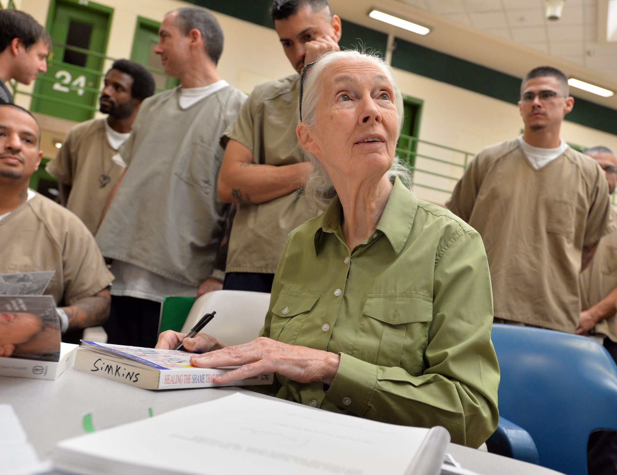 Jane Goodall signing a book titled "Healing the Shame That Binds You" while several men inmates stand nearby in the Boulder County Jail.