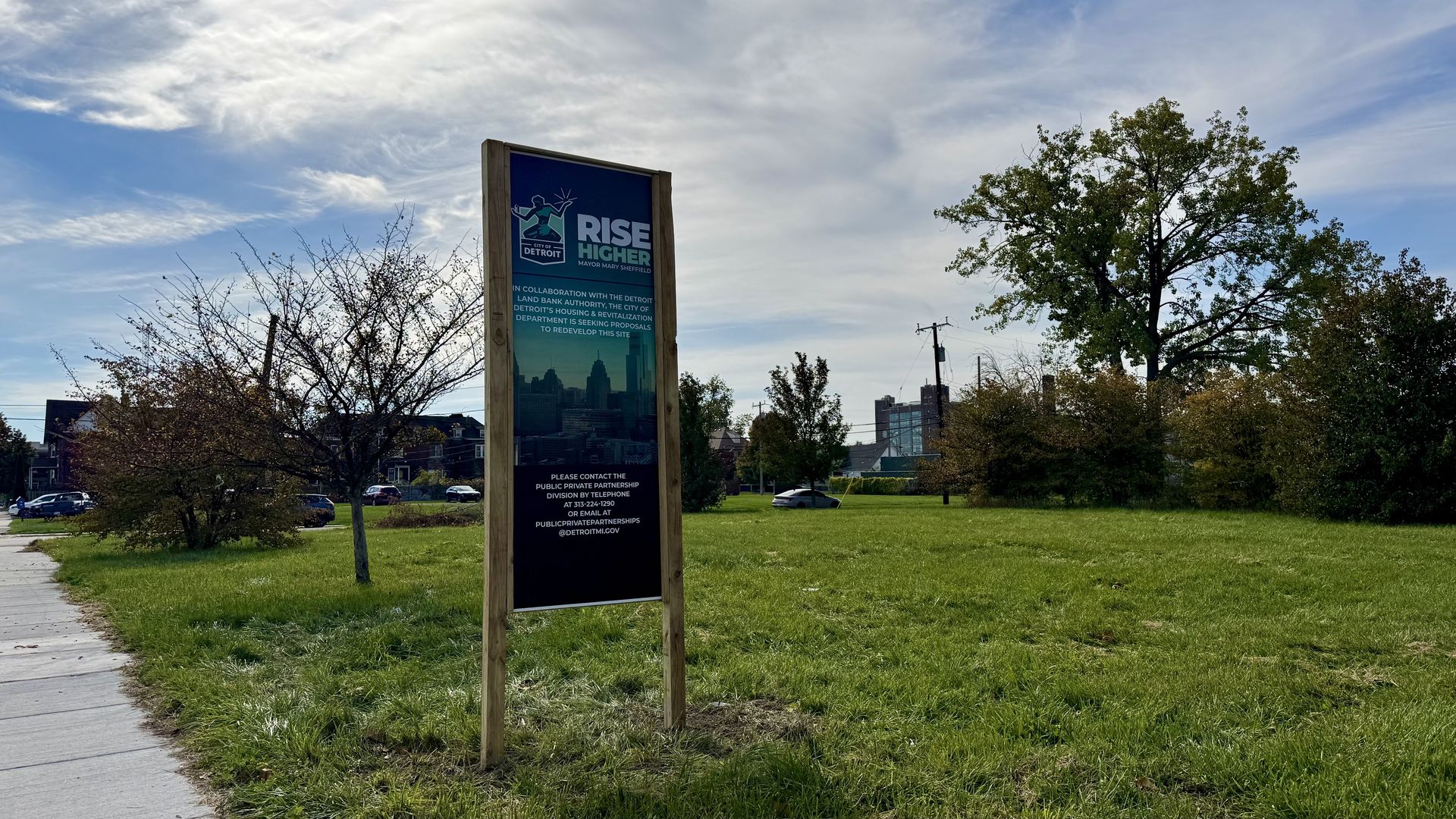 Tall wooden sign in a grassy park reads "RISE HIGHER" with the Detroit logo; distant city skyline visible, leafless shrubs left, a large leafy tree to the right, under a blue sky with clouds.