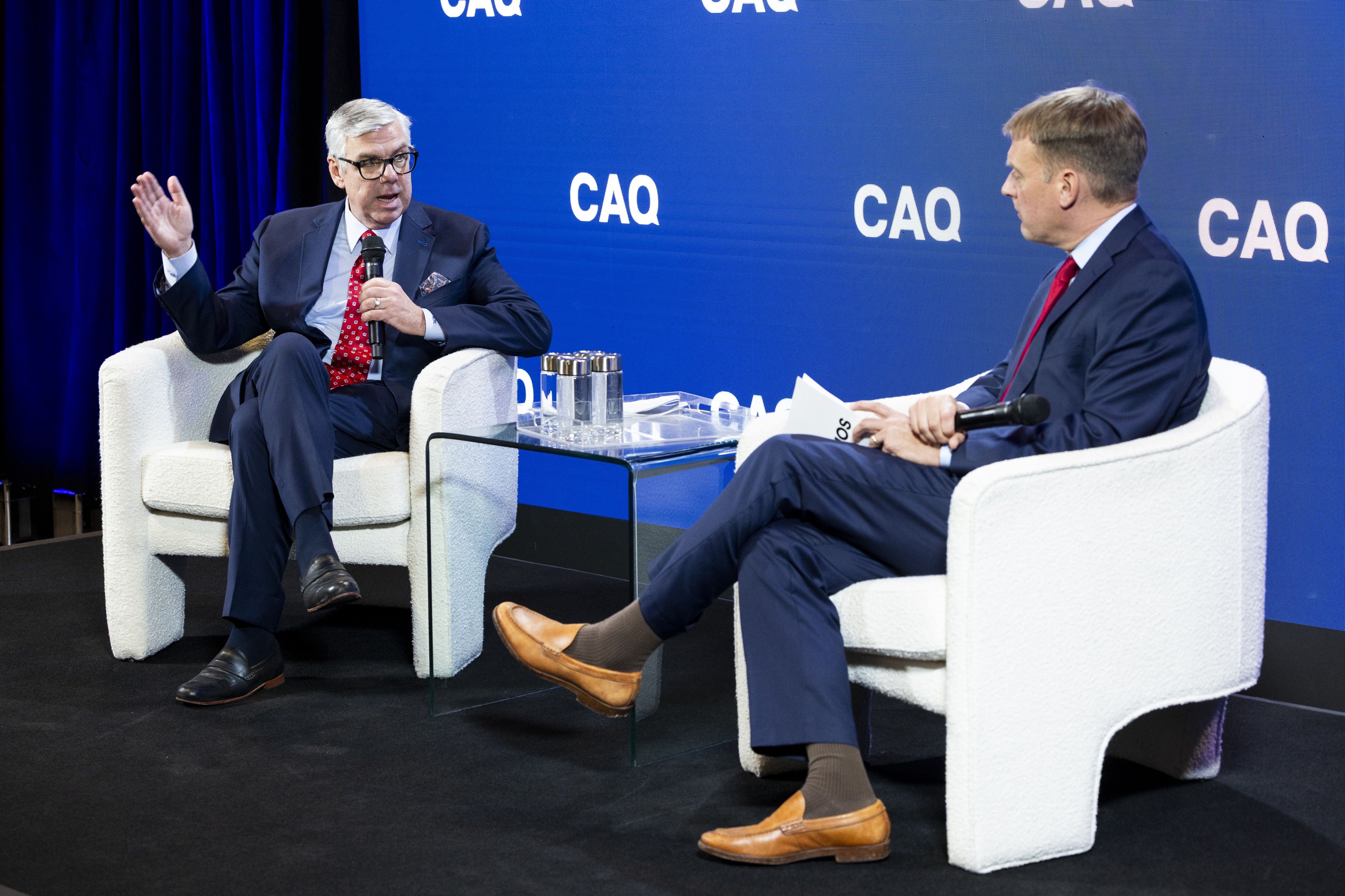 Two men in dark suits sit in white armchairs on a stage with a blue CAQ backdrop. The left man speaks into a microphone and gestures; the right man listens, holding papers.