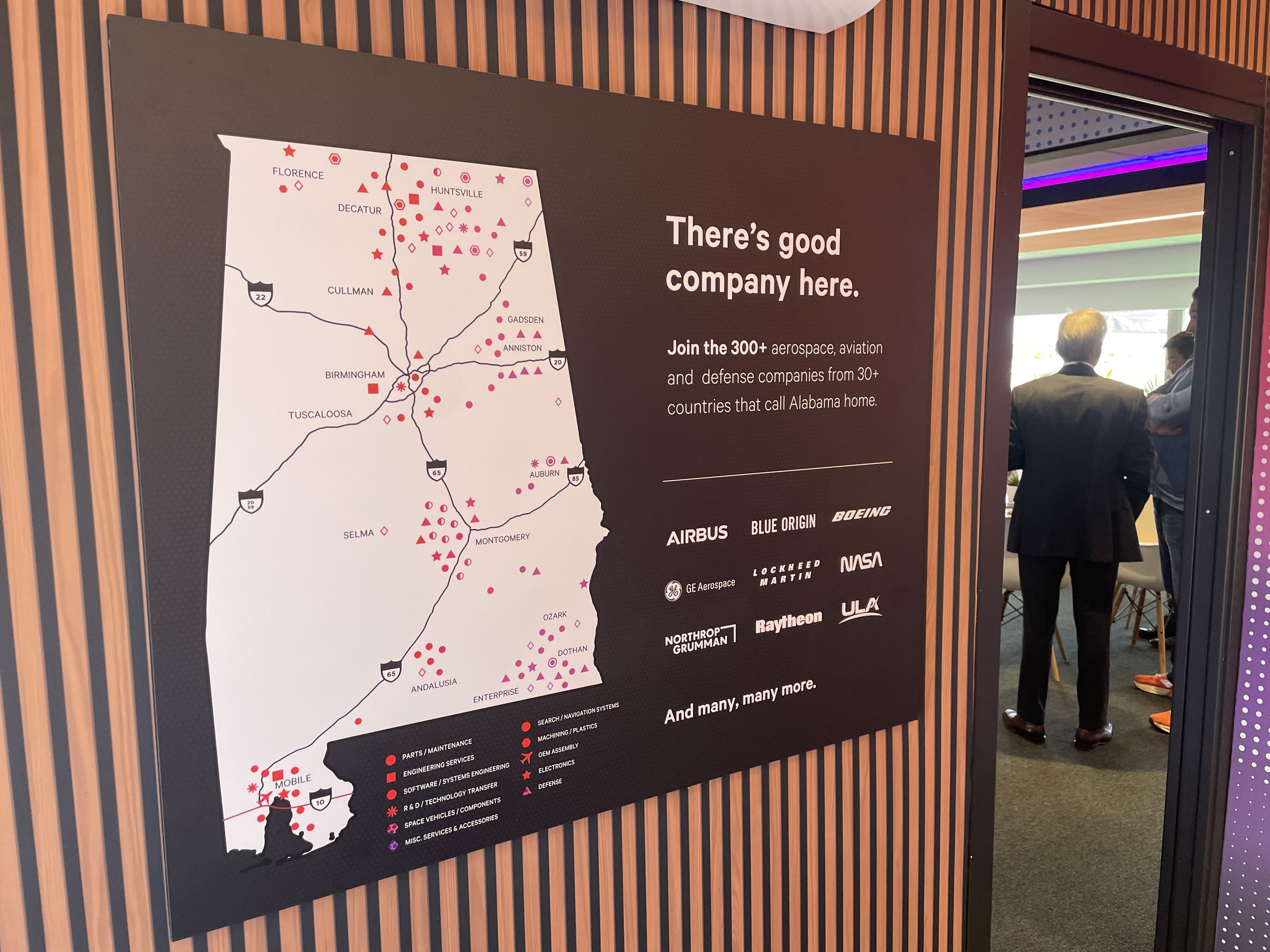 Large display board with a map of red icons on the left and a dark panel on the right that reads 'There's good company here.' Logos of aerospace companies appear below; people stand near an open doorway on the right.