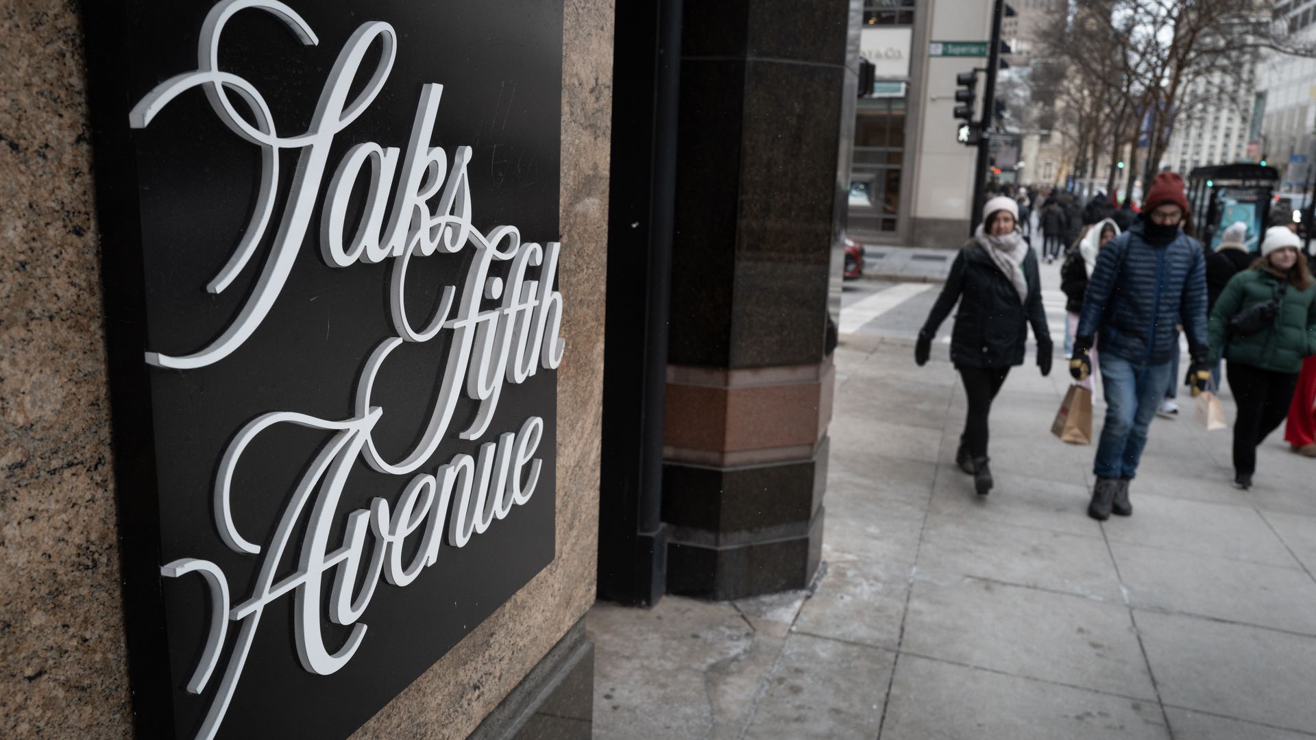 Close-up of a black plaque with white cursive sign reading "Saks Fifth Avenue" on a stone building; city street with pedestrians in winter coats in the background.