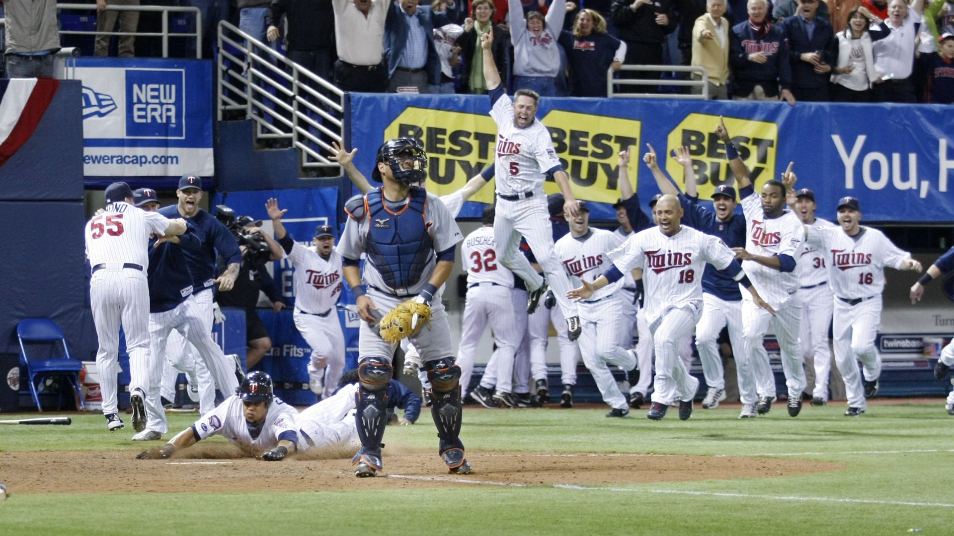 A baseball team wearing white pinstriped Minnesota Twins uniforms run out onto a baseball field as a teammate slides into home base with a catcher wearing the Detroit Tigers' gray uniforms stands in the foreground.