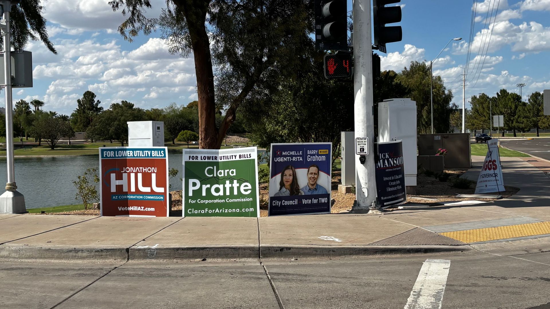 Bright street corner with a red traffic light on a tall pole, campaign posters along the sidewalk; a waterway and trees in the background under a blue sky with white clouds.