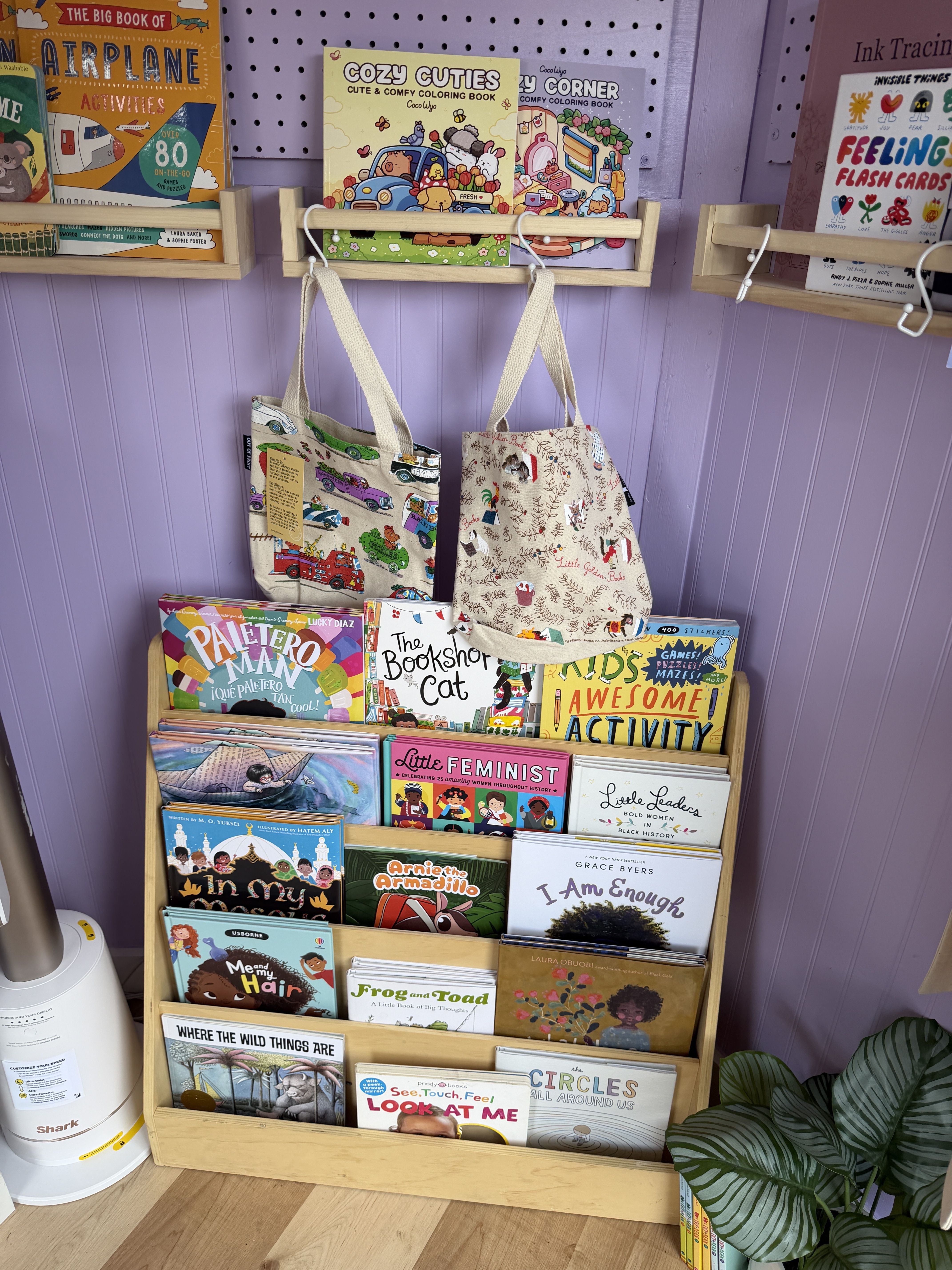 A lavender bookstore display with wooden racks and pegboard, two beige tote bags hanging, and colorful children's books like "Airplane Activities" and "Cozy Cuties".