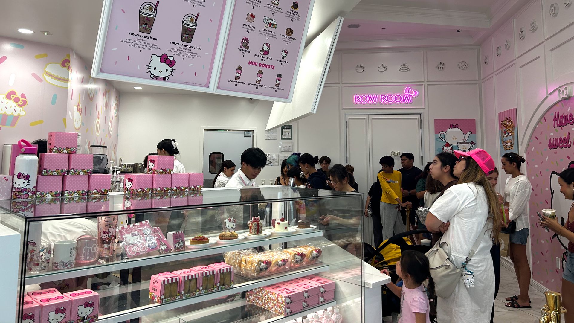 Inside a pink Hello Kitty-themed cafe with customers lining up, pink boxes, dessert display cases, and large menu boards featuring seasonal specials and desserts. Neon sign reads "BOW ROOM."