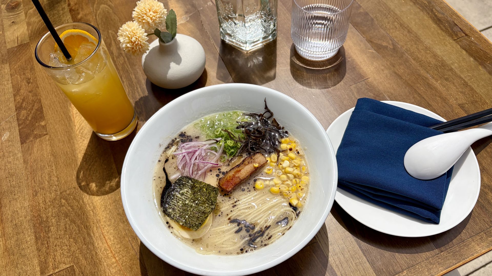 Top-down view of a ramen bowl with corn, seaweed, pork, and onions on a wooden table; a glass of orange juice with ice and straw; a small vase with pale flowers; a water glass; and a navy napkin with chopsticks.