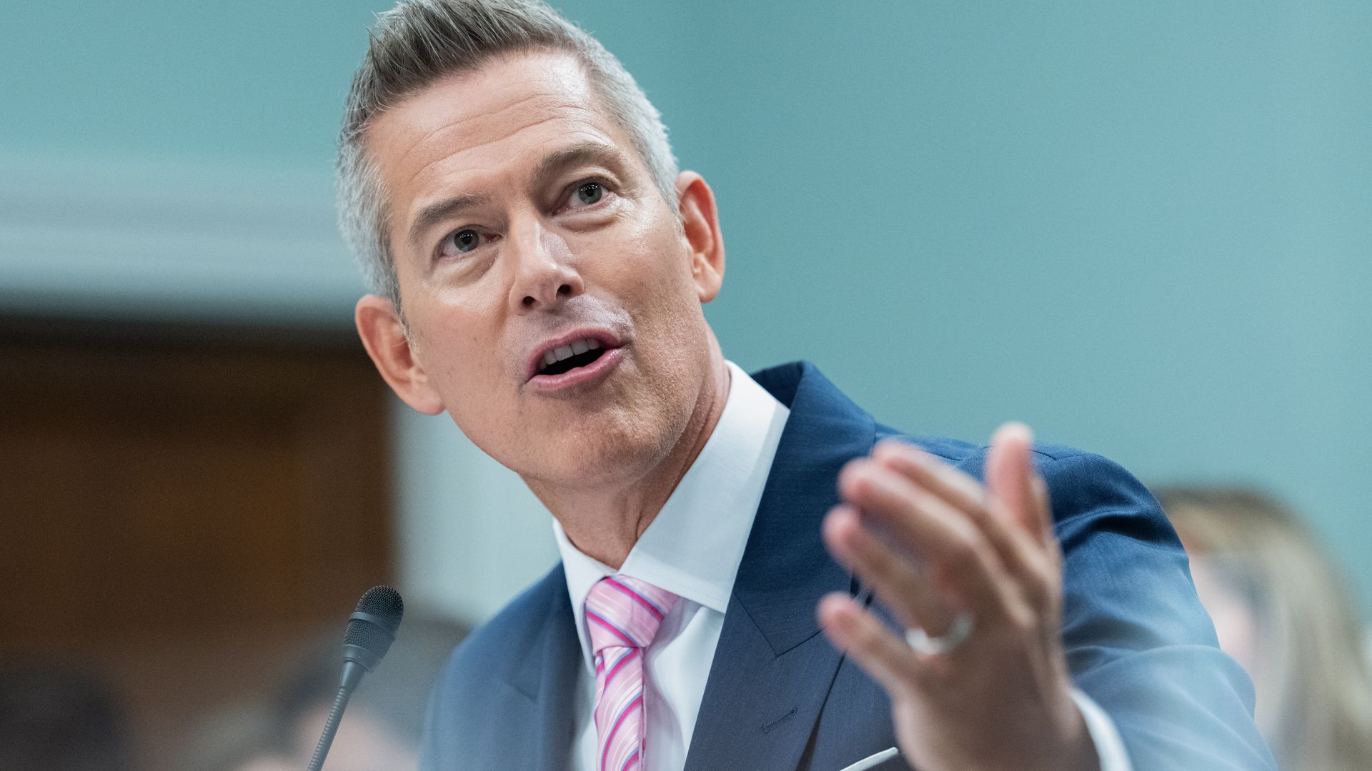A man with short salt-and-pepper hair and wearing a navy suit and pink striped tie gestures toward the camera while testifying in a government setting.