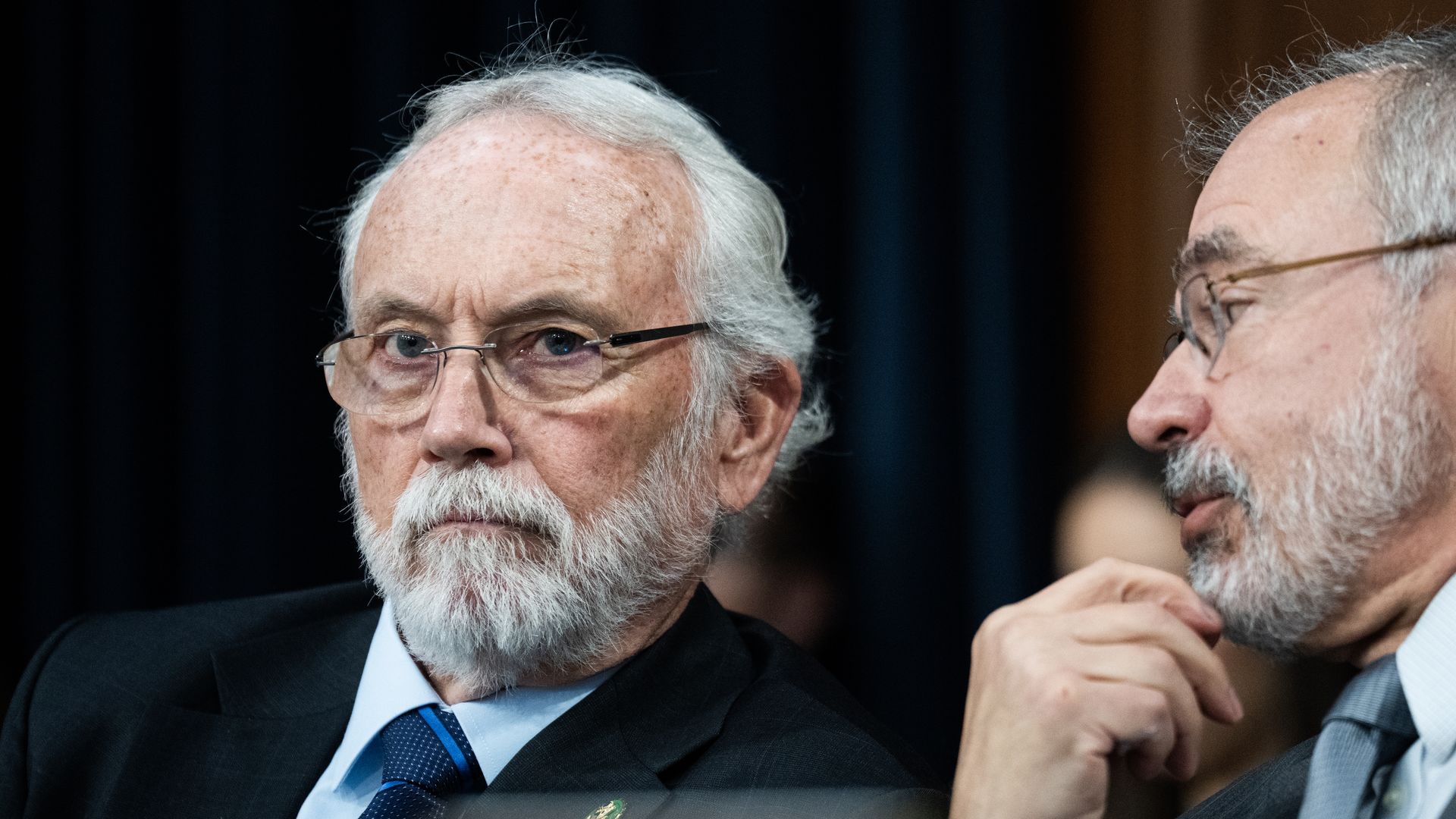 Two men with gray hair and beards wear glasses and suits, sitting closely. The man on the left looks serious, wearing a blue tie, while the man on the right appears to speak softly.