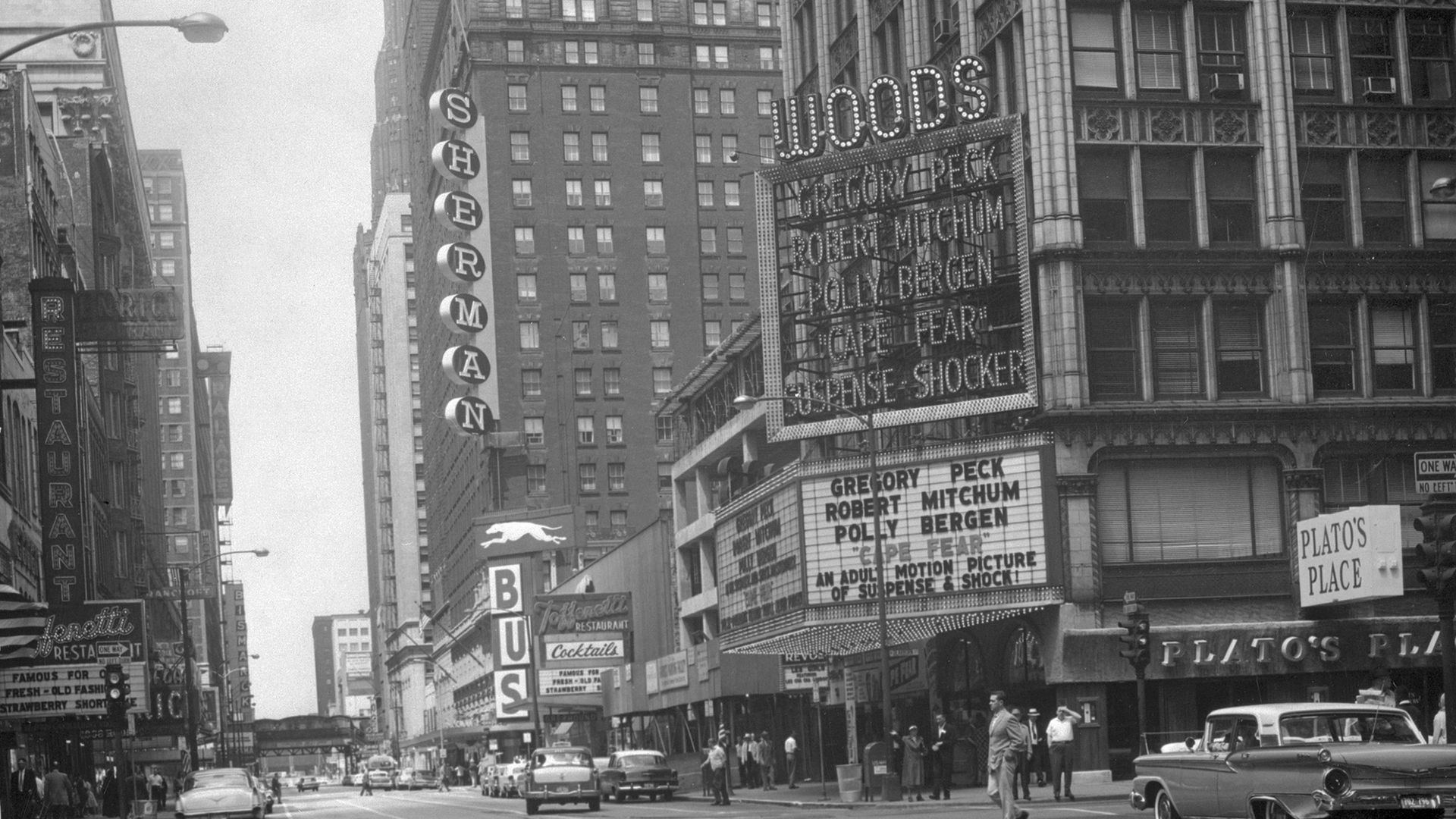 Photo of a street with big signs like "BUS" and WOODS" along a street in the 1960s. 