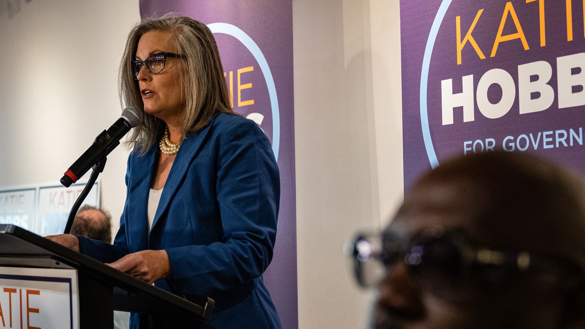 A woman speaks into a microphone in front of signs that say Katie Hobbs for governor.