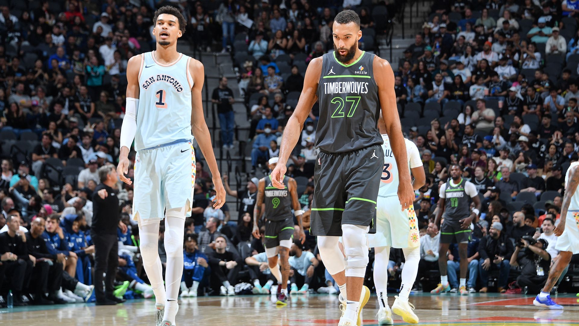 Two basketball players walk on a crowded court during a game; one in a light blue San Antonio jersey (#1) and the other in a dark Timberwolves jersey (#27), with fans in the stands.