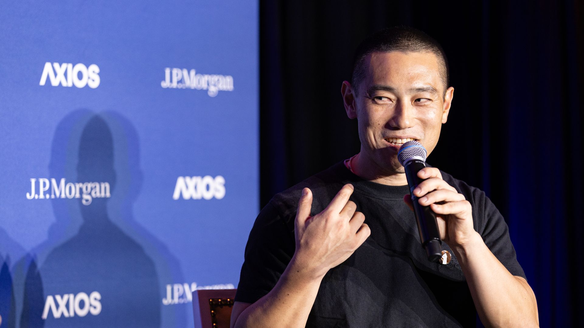 Smiling man in a black shirt speaks into a microphone on stage. A blue backdrop displays AXIOS and J.P. Morgan logos, with dark curtains visible on the right.