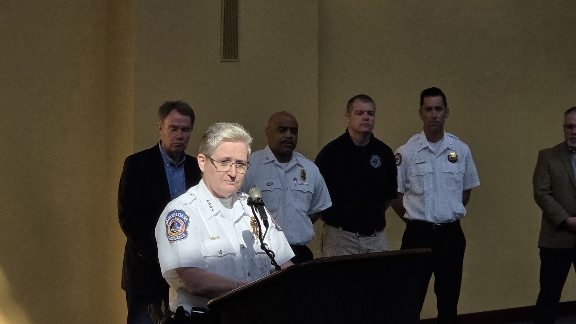 A female police officer in a white uniform speaks at a podium with a microphone, flanked by several officers in light blue shirts in a beige conference room.