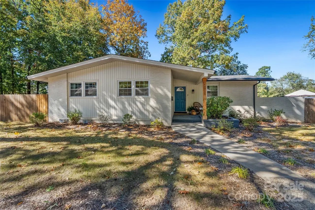 Single-story white house with a teal front door, surrounded by trees with fall foliage, a fenced yard, and a concrete walkway leading to the entrance under a clear blue sky.