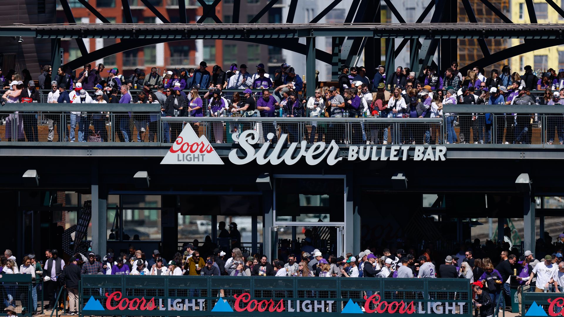 Fans in purple stand behind a Silver Bullet Bar sign wearing purple. There's ballpark beams behind them. 