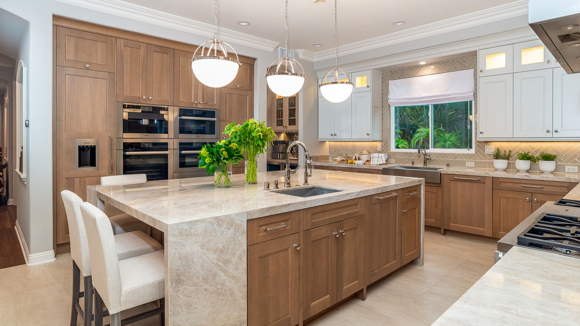 Bright modern kitchen with a large marble island, brown wood cabinets, white upper cabinets, and stainless steel double ovens. Three pendant lights over the island; a window shows greenery outside.