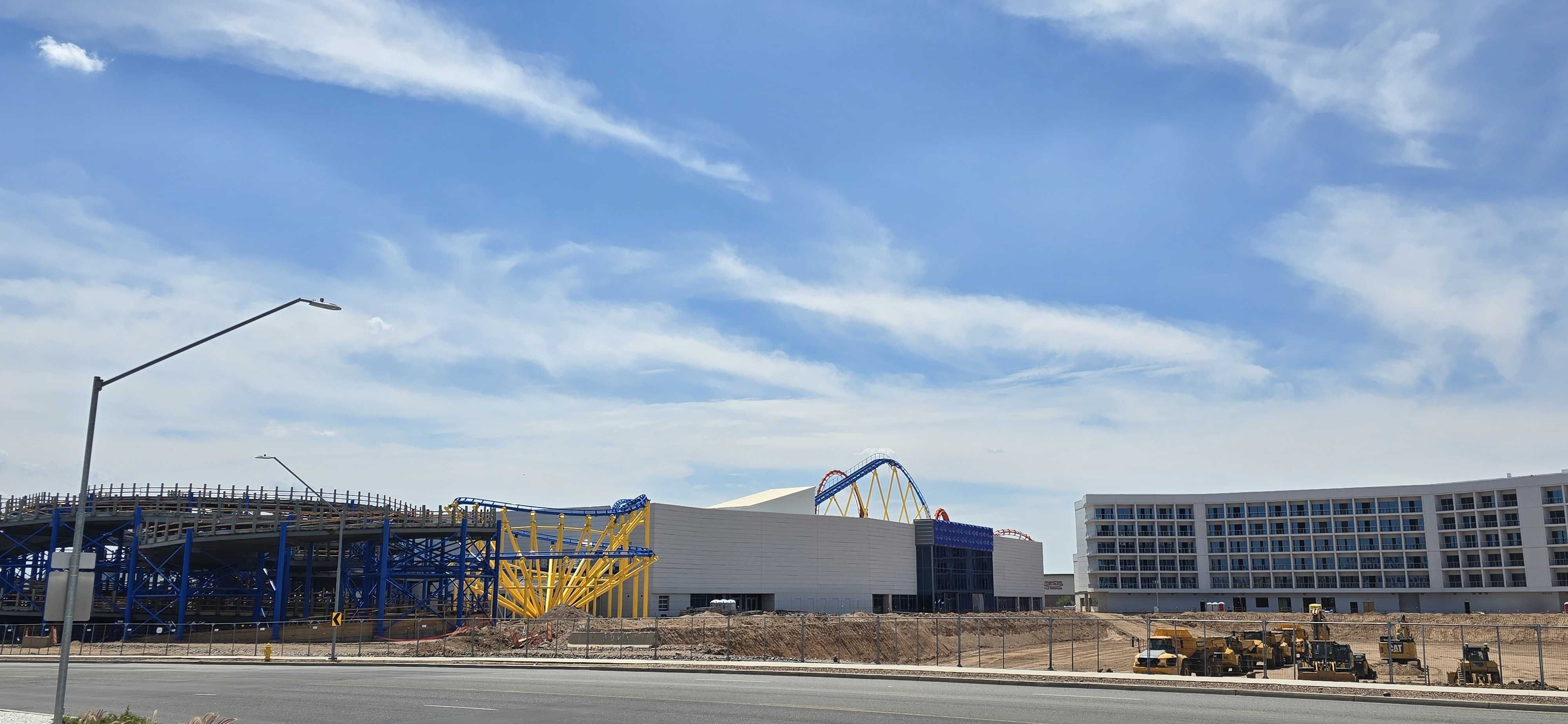 Wide view of a construction site beside a modern building under a bright blue sky with wispy clouds. A blue and yellow roller coaster frame rises among fencing and heavy equipment in the foreground.