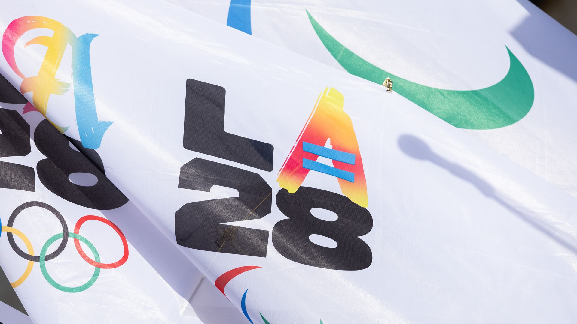 Close-up of a white fabric with colorful printed text "LA 28" and Olympic rings, with a green Nike swoosh logo partially visible in the background.