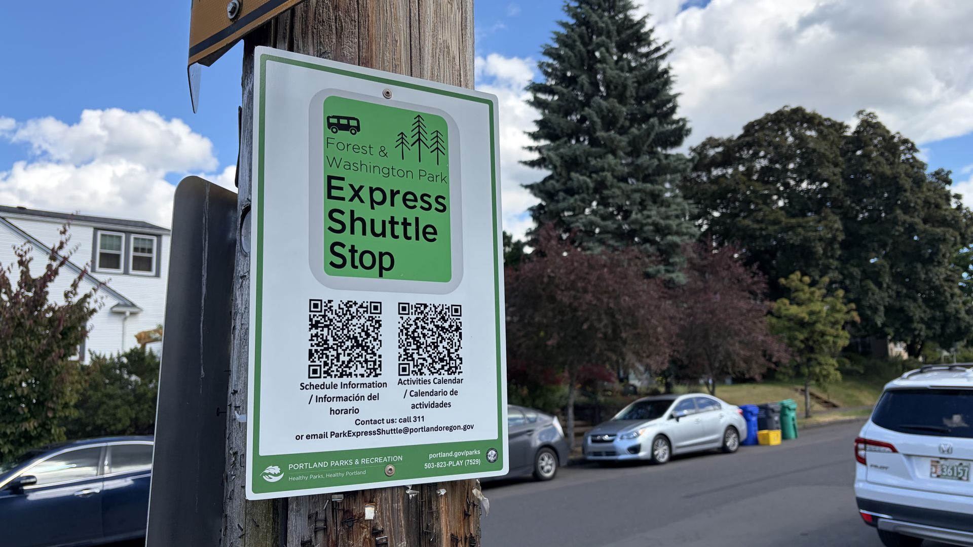 Green and white sign on a weathered wooden post reads Forest & Washington Park Express Shuttle Stop, with a bus icon and trees; a QR code sits at the bottom as cars line the street.