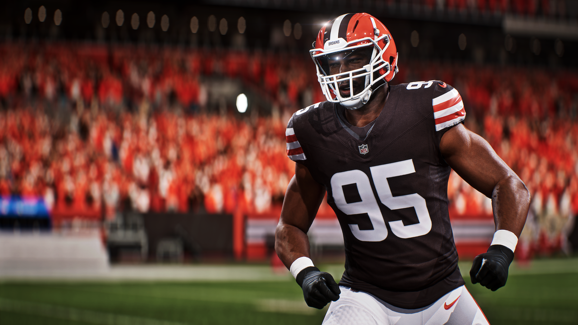 NFL player in black jersey number 95 with orange and white striped helmet and gloves, running on a football field with a blurred crowd in red in the background.