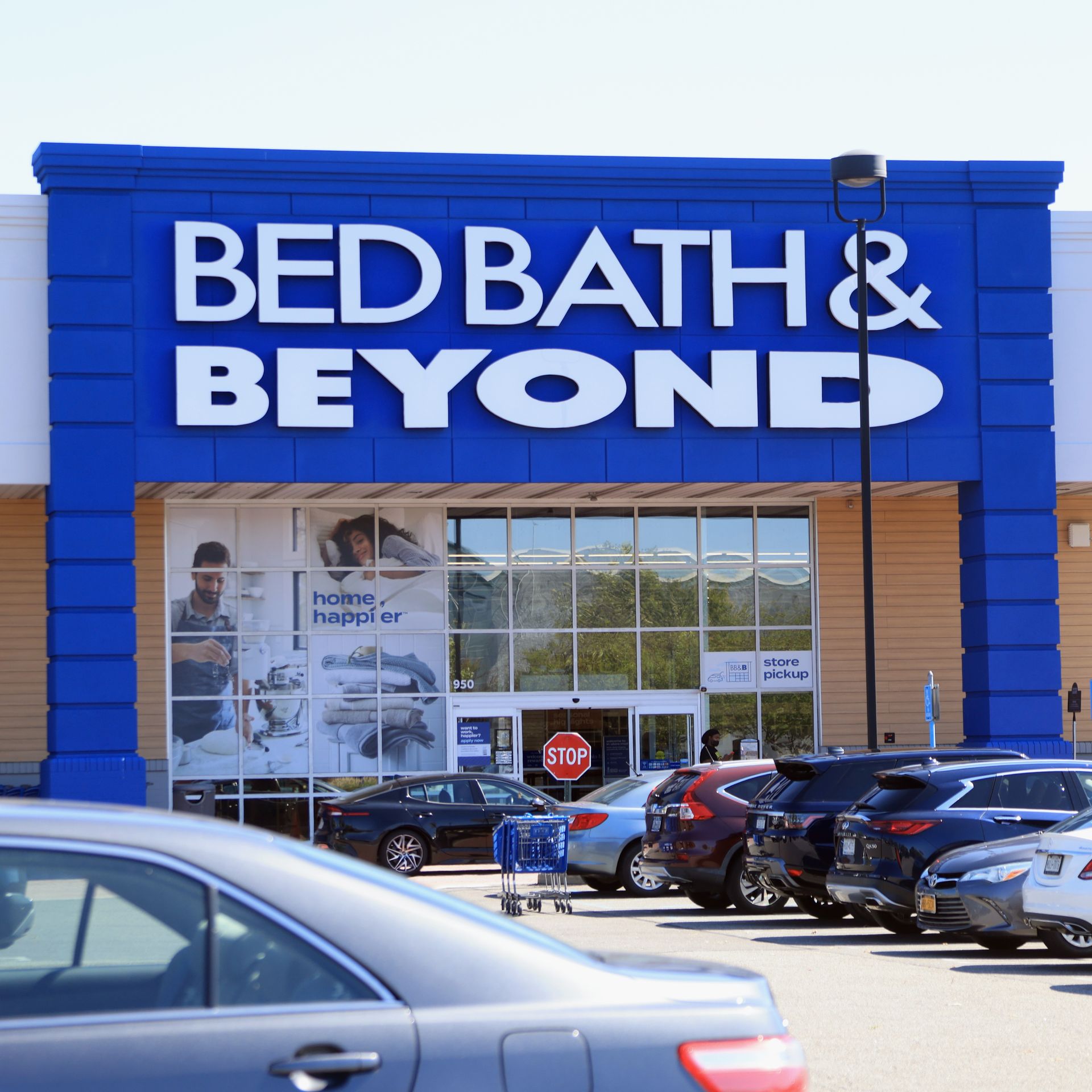 A Bed Bath & Beyond store front, in blue and white, with a parking lot filled with cars.