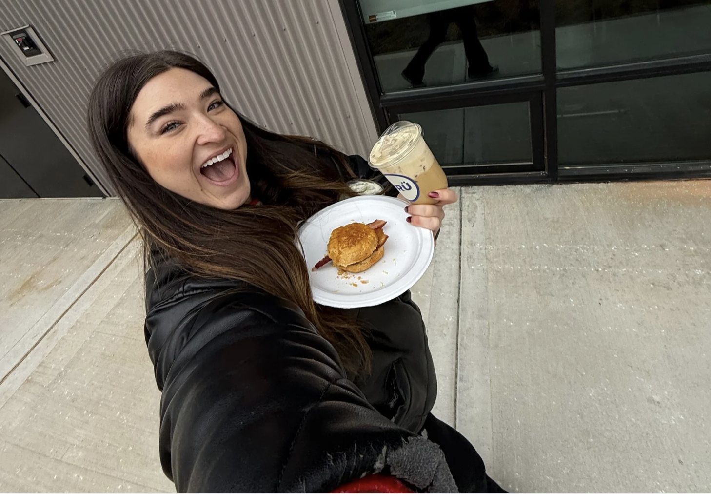 Smiling woman with long brown hair in a black jacket holds a plate with a sandwich and a cup of iced coffee outside on a concrete sidewalk near a building with glass windows.