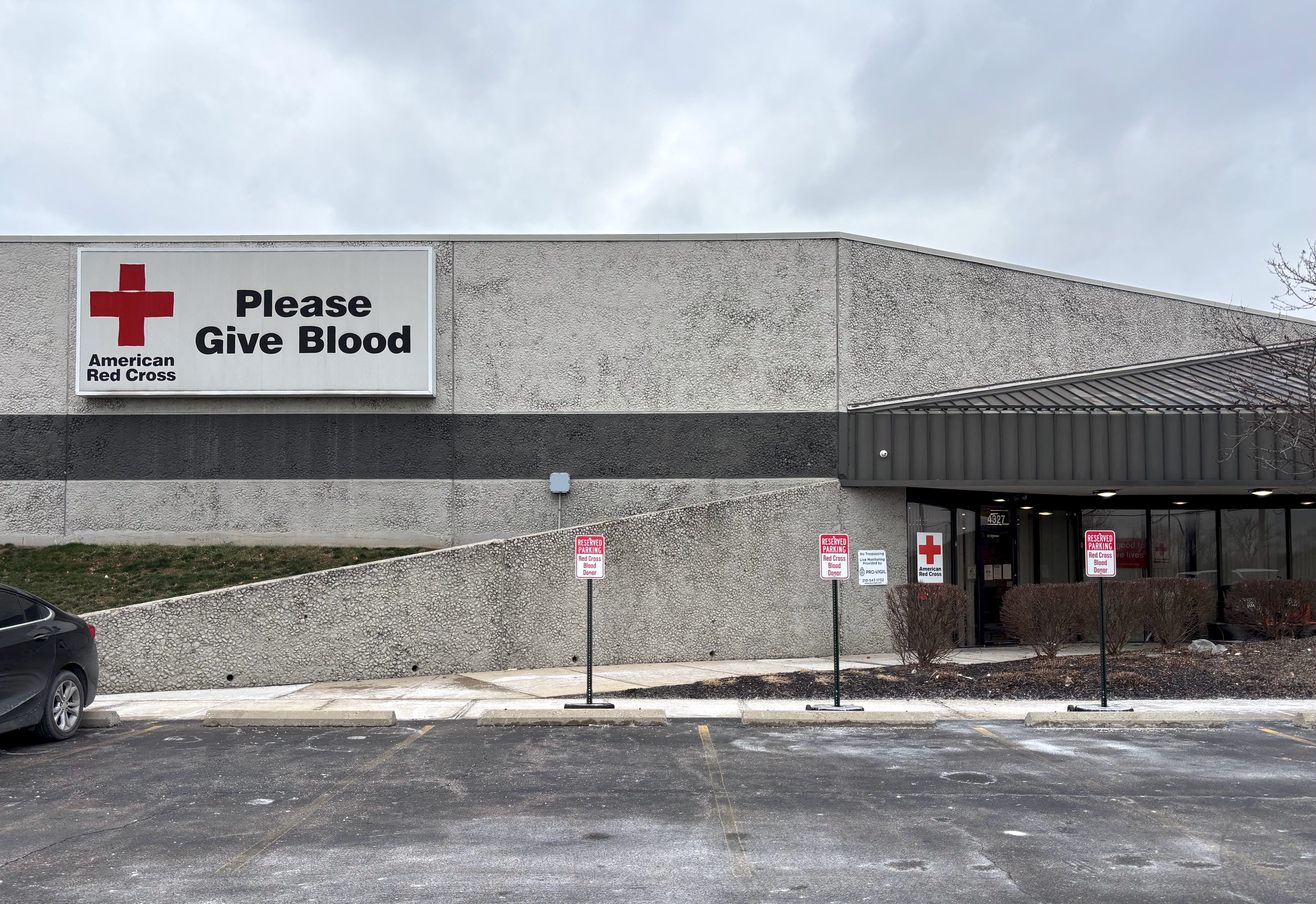 Red Cross donation center with large sign reading "Please Give Blood" and American Red Cross logo, reserved parking signs for blood donors in front, under cloudy sky.