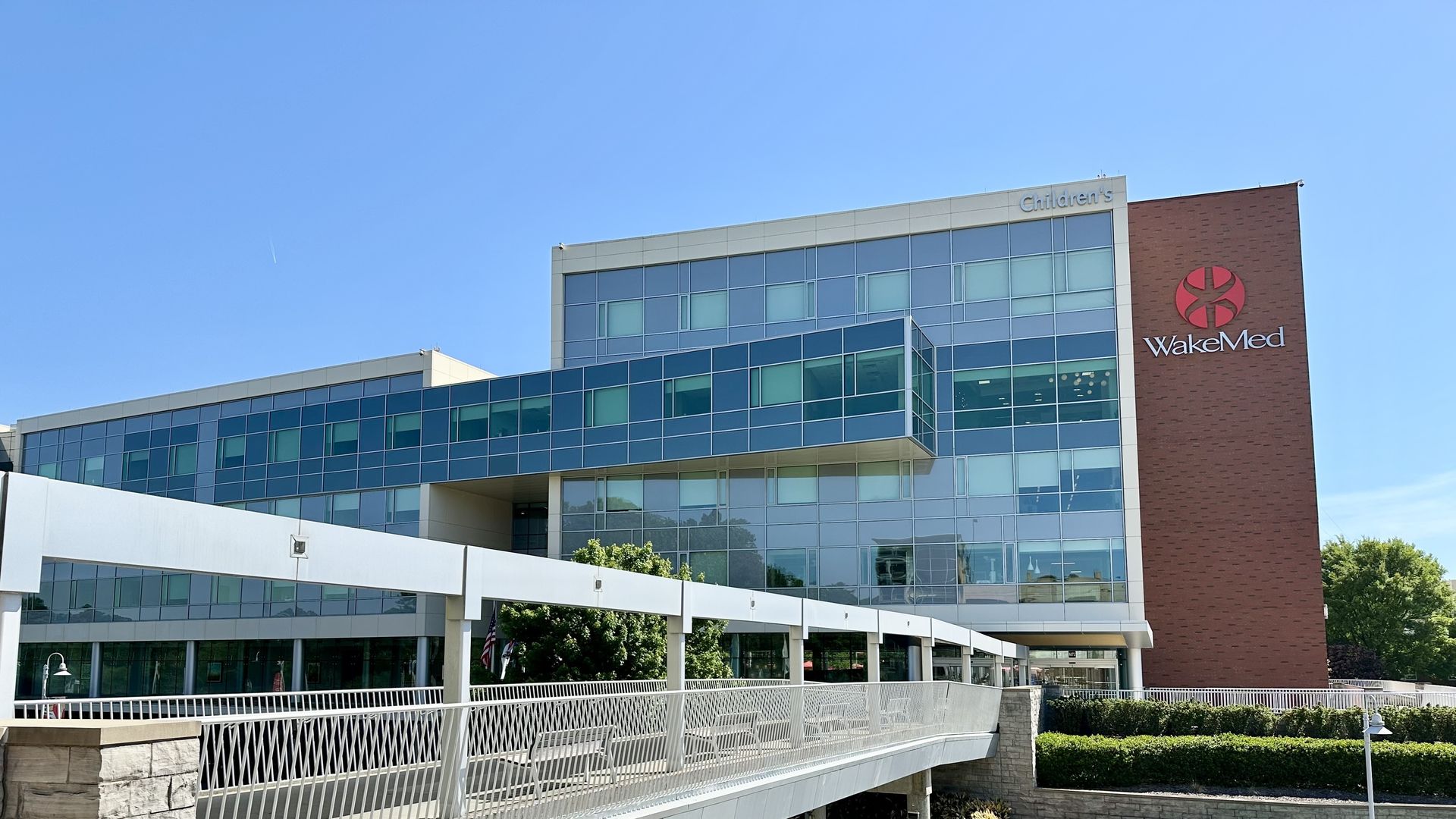Modern glass-front hospital building with a red brick tower bearing the WakeMed logo. A Children's sign atop the building; a white elevated pedestrian bridge crosses the foreground under a blue sky.