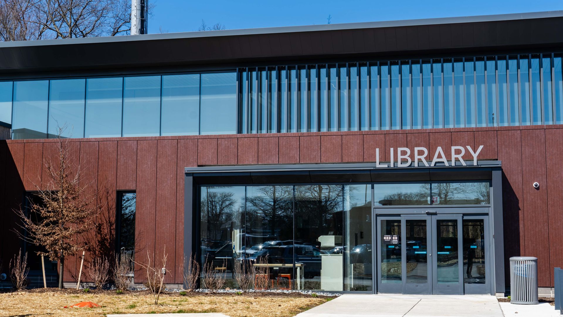 Modern library building with large glass windows, a red brick facade, and double doors. The word LIBRARY is in white above the entrance, with leafless trees and a blue sky behind.