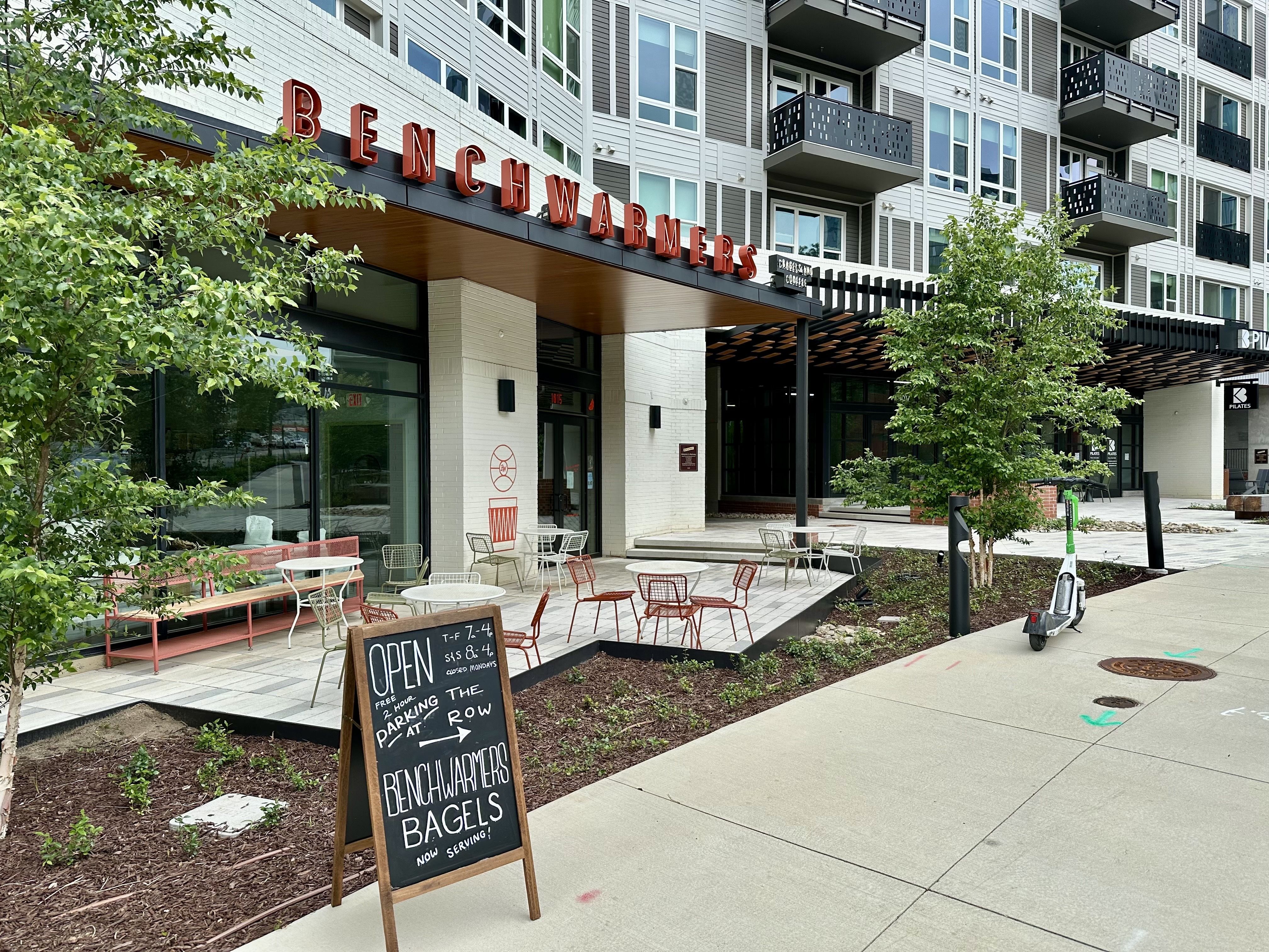Wide view of a storefront with a red "BENCHWARMERS" sign, outdoor seating with red and white chairs, chalkboard signs, and a modern apartment building with balconies; trees line the sidewalk.