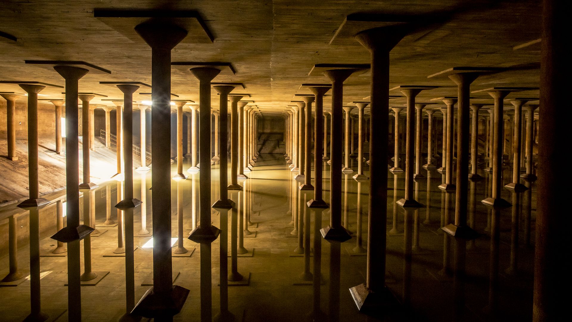 Long underground hall with evenly spaced concrete pillars rising from shallow water, warm lights along the left cast reflections on the water, leading to a distant vanishing point.