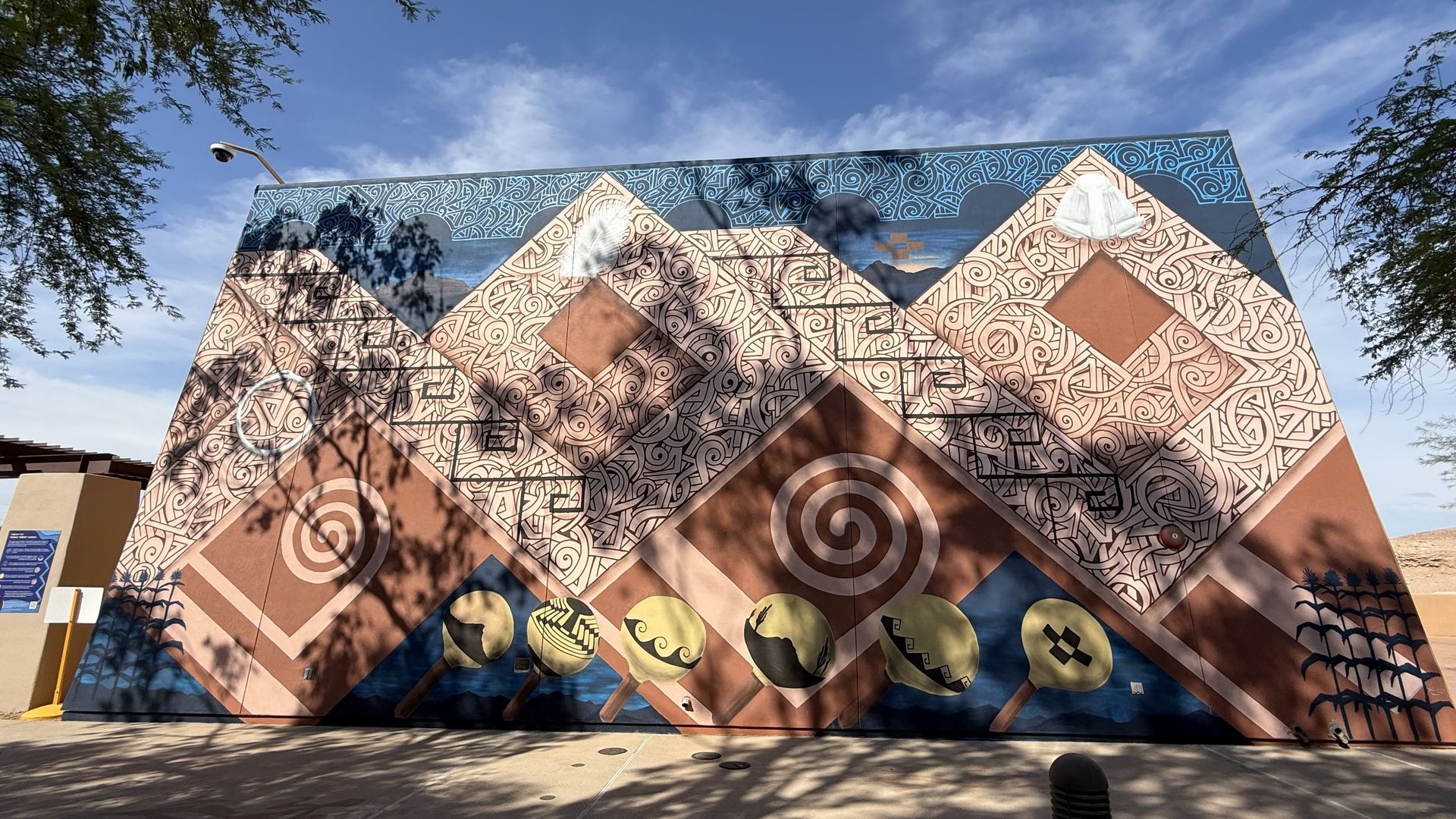 Colorful mural on a triangular building featuring beige spiral patterns forming mountains, a blue patterned top, and yellow circular emblems at the base; tree shadows cross the wall under a blue sky.