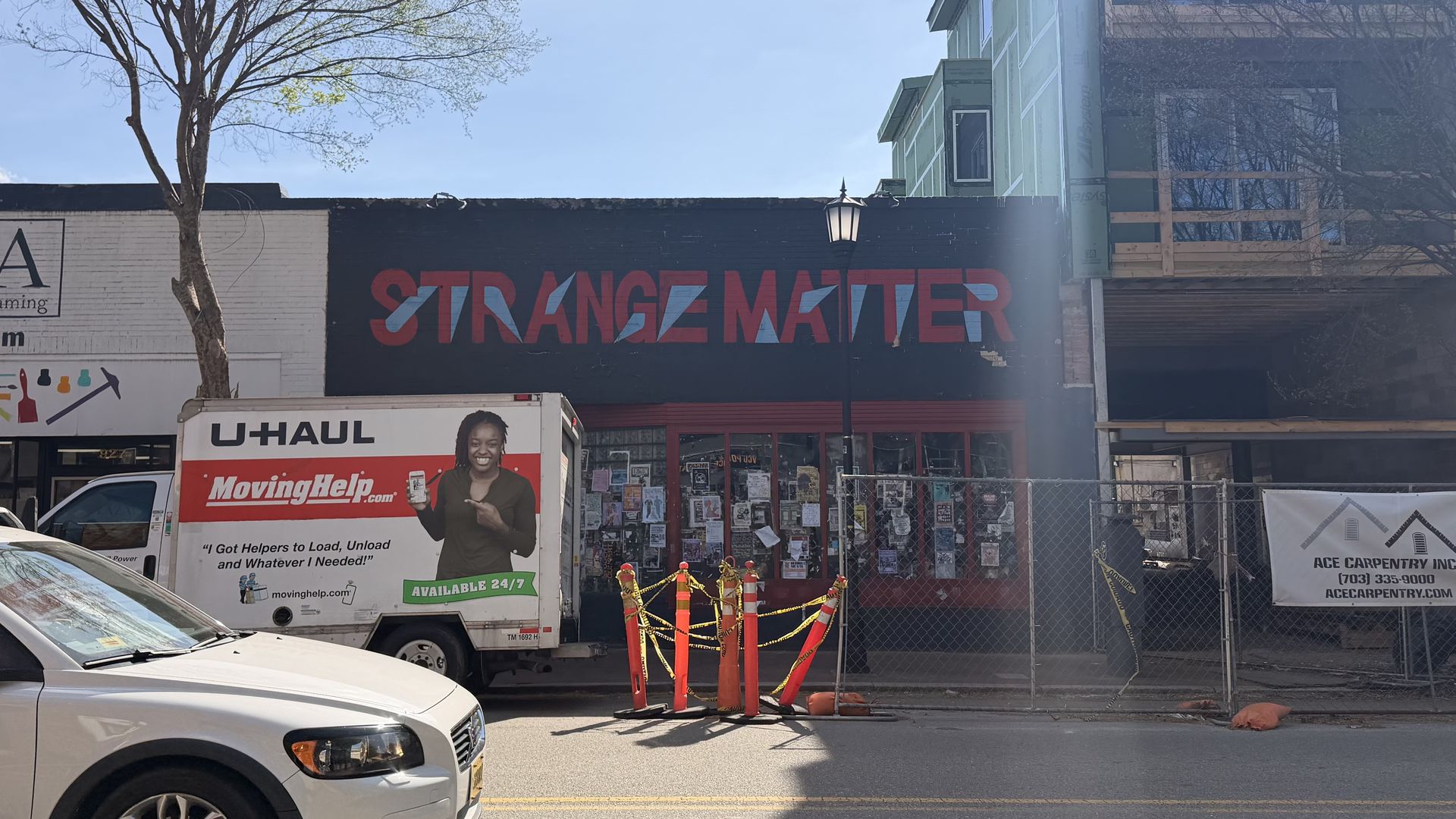 Street scene with storefronts: left PLAZA Art Materials, center "STRANGE MATTER" mural in red and blue, U-Haul truck and white car parked, orange cones, blue sky, bare trees, and a building under construction on the right.