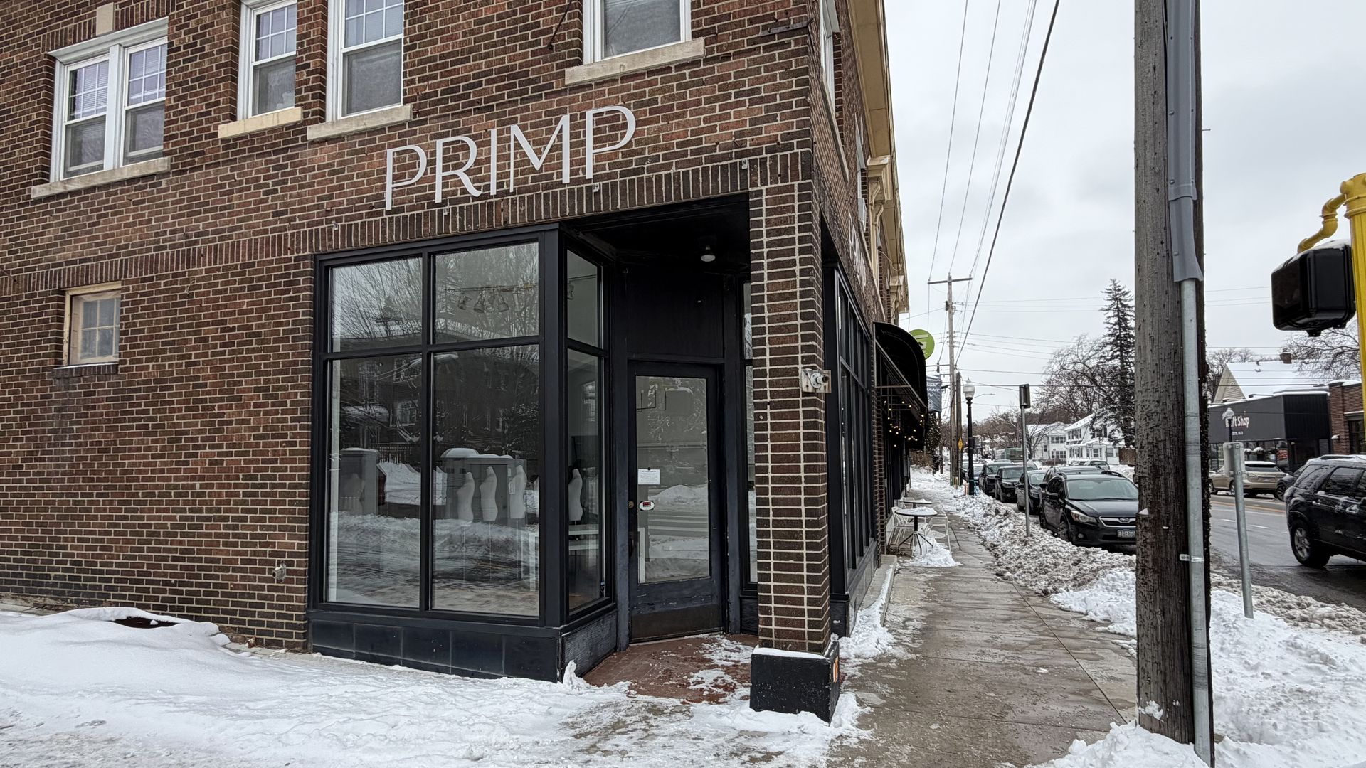 Brick building corner with a black-framed glass door and windows labeled "PRIMP", snowy sidewalk and street with parked cars under a cloudy sky.
