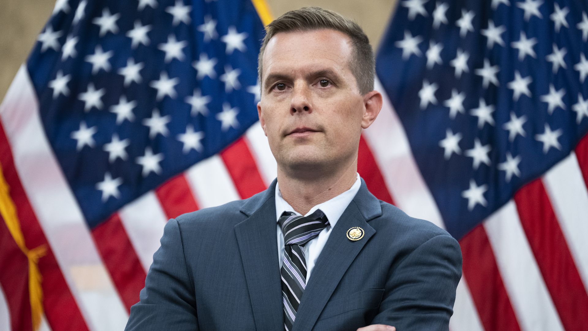 Portrait of a man in a blue suit and striped tie, standing with arms crossed in front of American flags; lapel pin visible, serious expression.