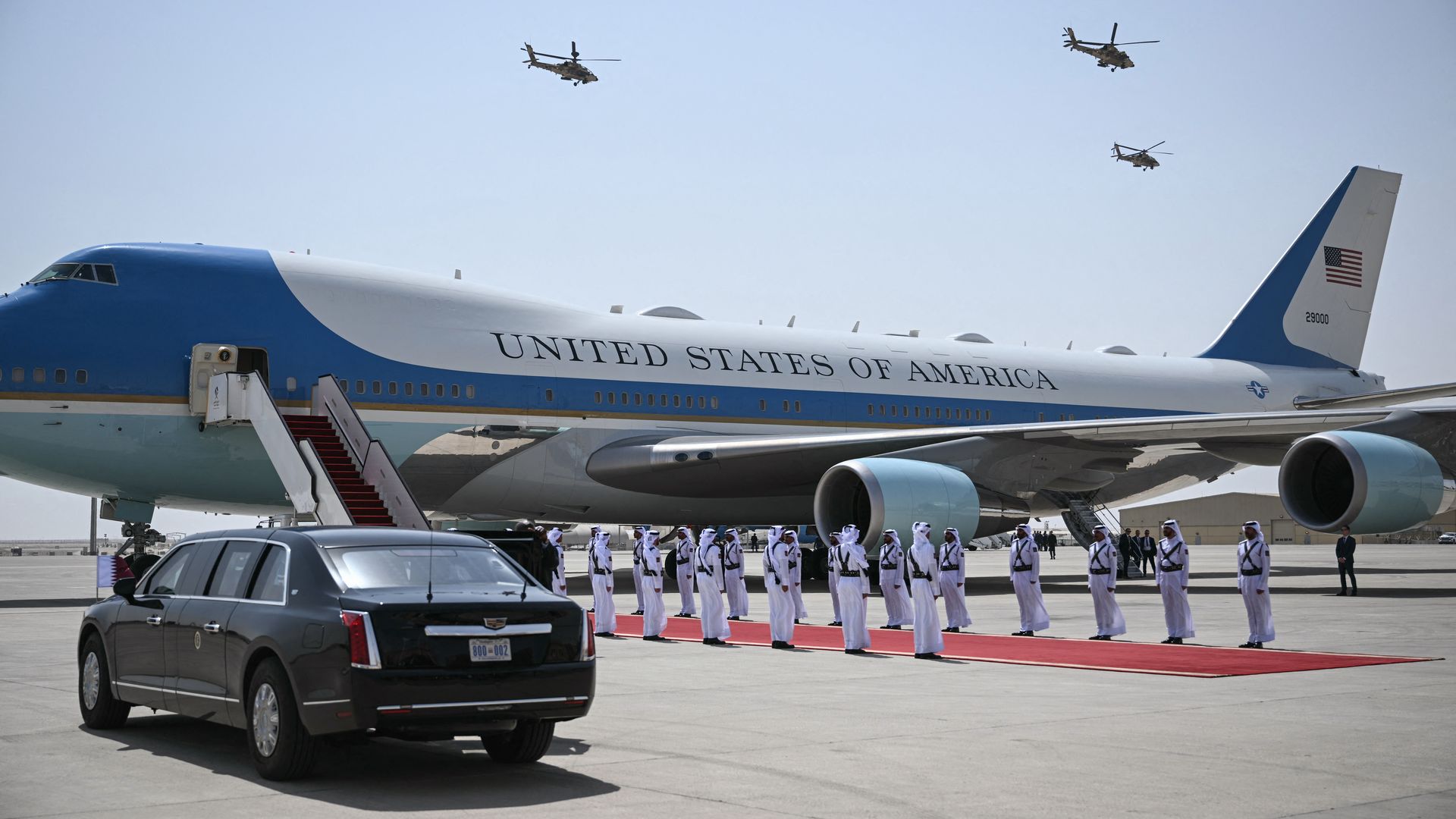Air Force One sits on tarmac, with a large black vehicle parked near the front. Helicopters fly overhead. People stand near a red carpet near the airplane's stairs.