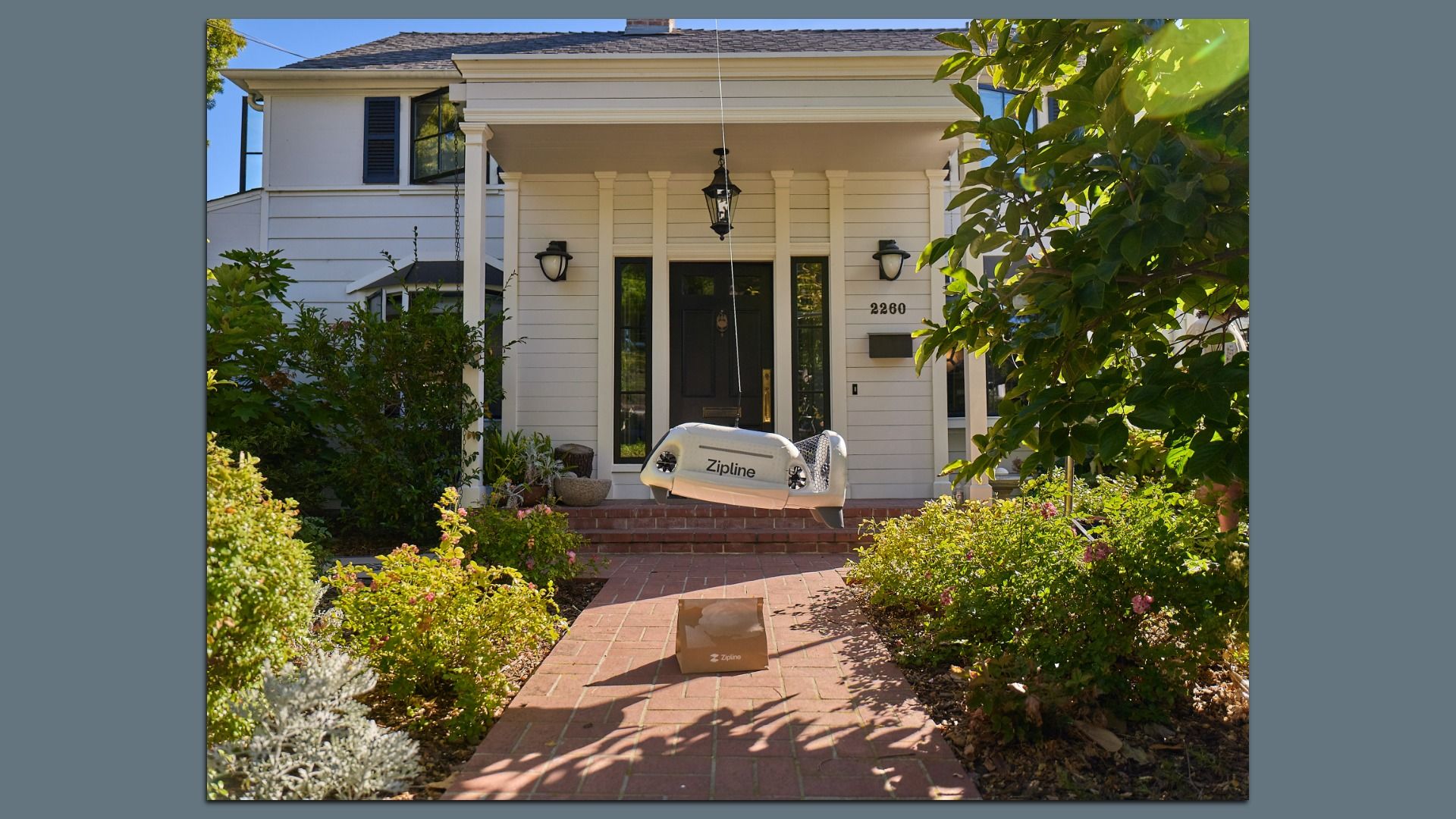 Front yard of a white house with black door, a drone with "Zipline" written on it hovers above a brick pathway delivering a package, surrounded by green bushes and sunlight.