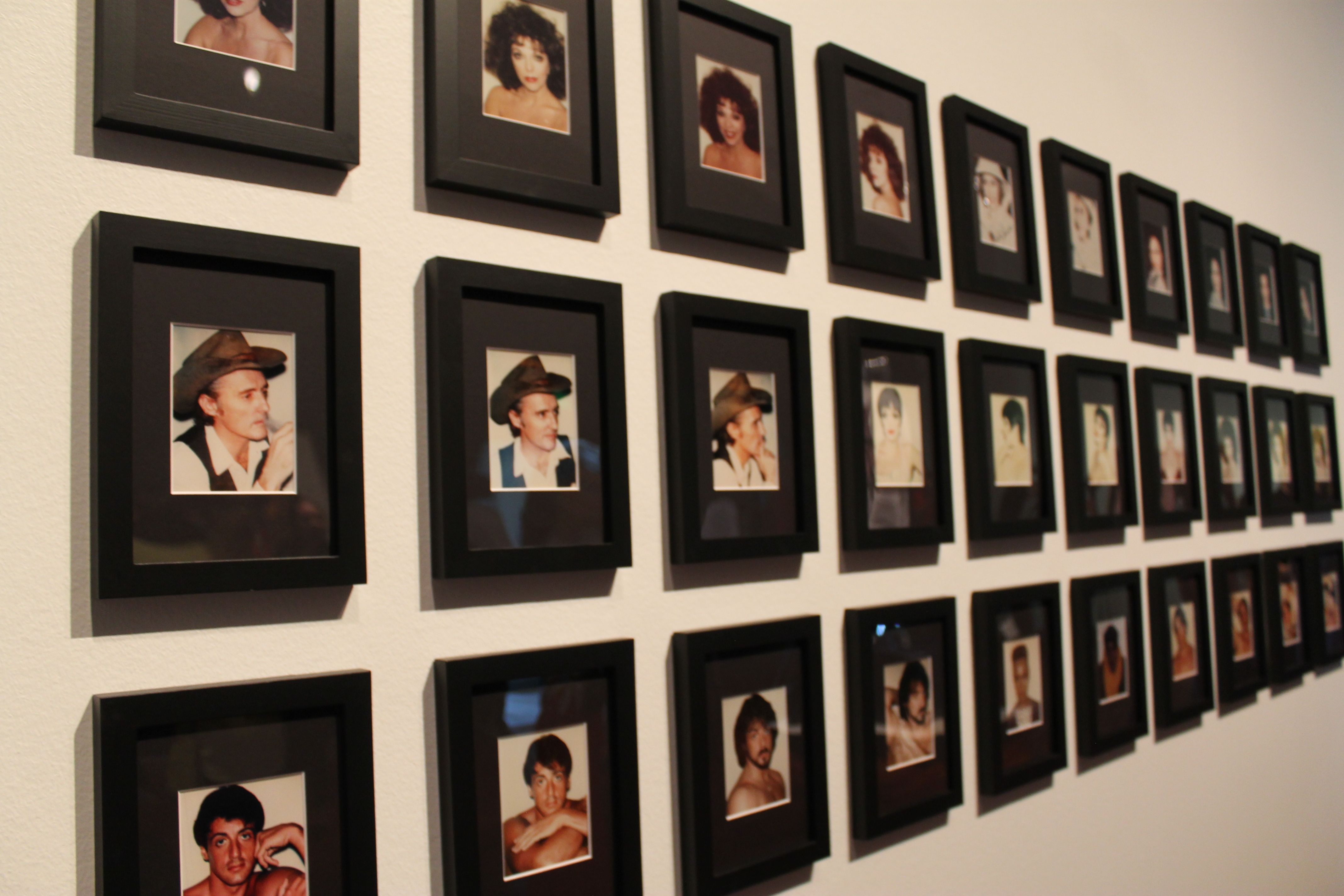 A gallery wall of many small black-framed portraits arranged in a grid on a pale wall, showing vintage headshots of men and women; some wear hats or bold outfits.
