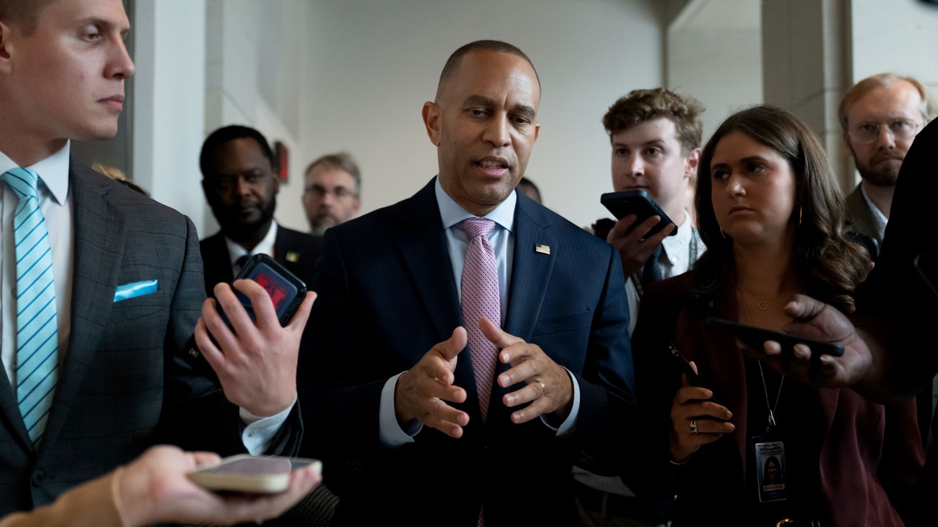Hakeem Jeffries, wearing a blue suit and speaking to reporters in a white and sandstone hallway while flanked by aides and security.